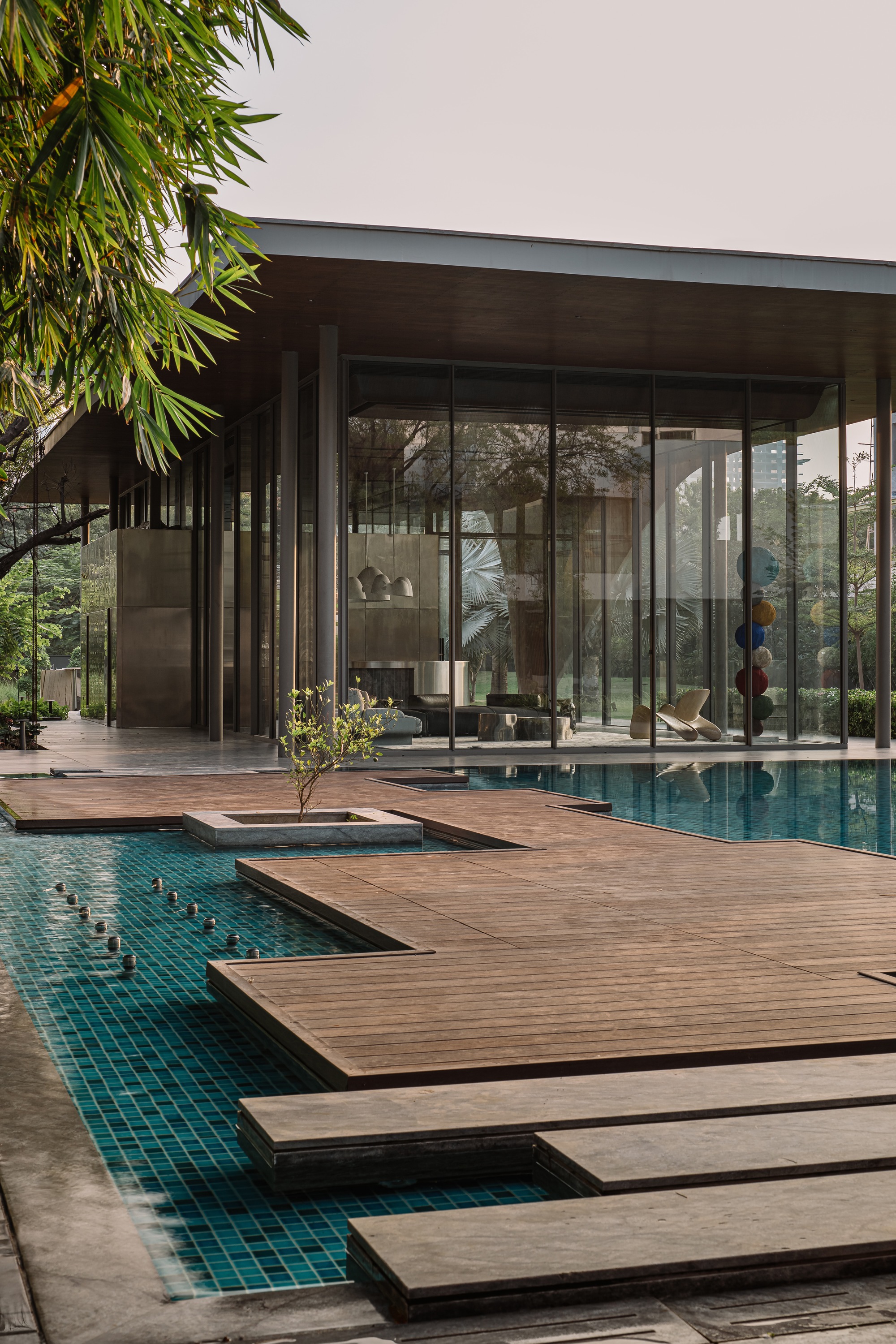 Stepped timber decking crossing the pool beneath a glass-walled pavilion with overhanging roof and bamboo above
