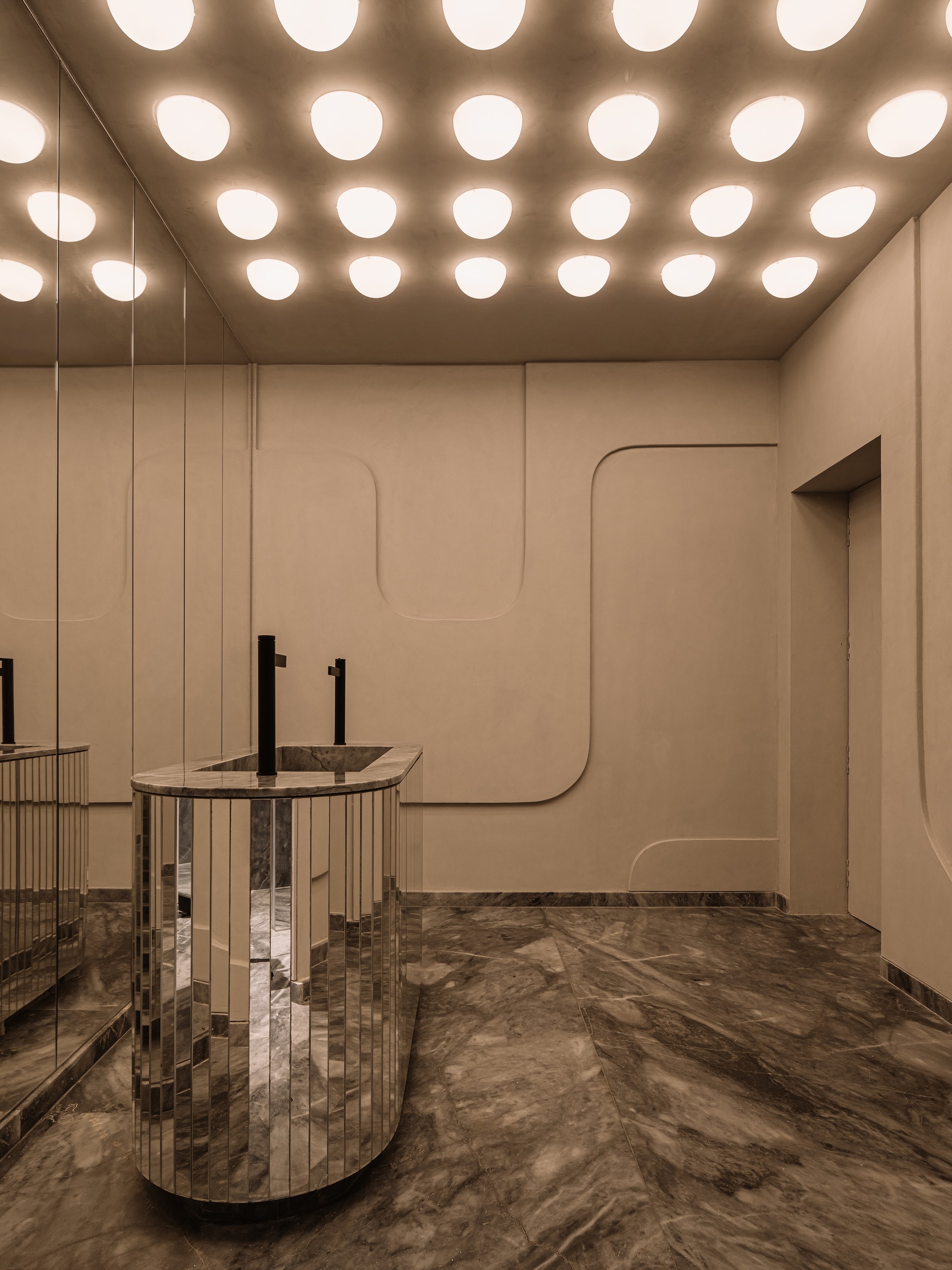 Bathroom with circular recessed ceiling lights above cylindrical mirrored sinks and sculpted wall panels in warm stone