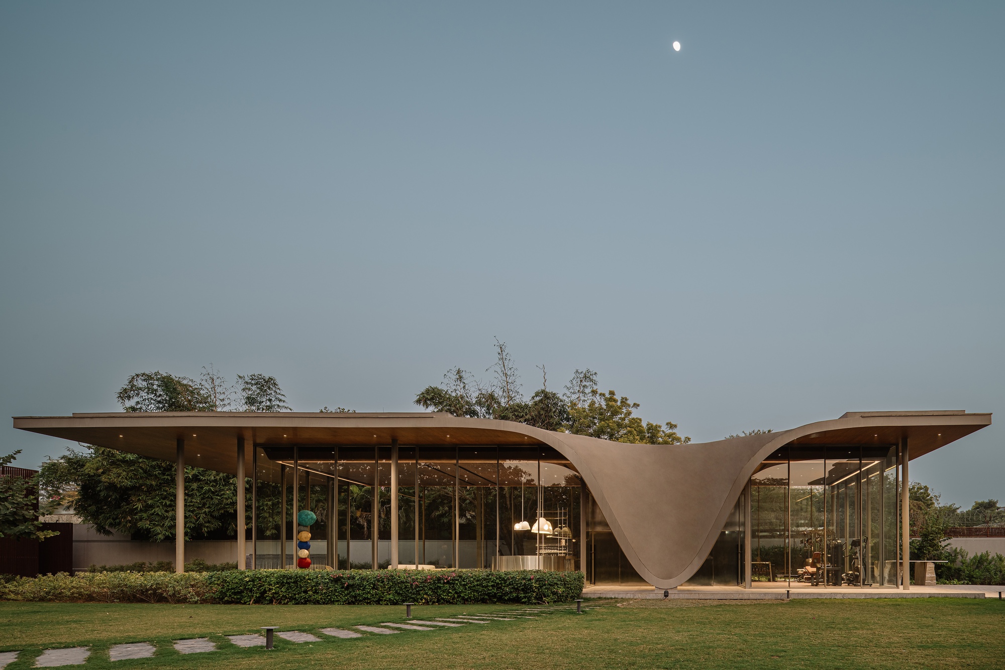 Glass pavilion with hyperbolic paraboloid roof connecting two volumes across a stepped stone pathway and lawn