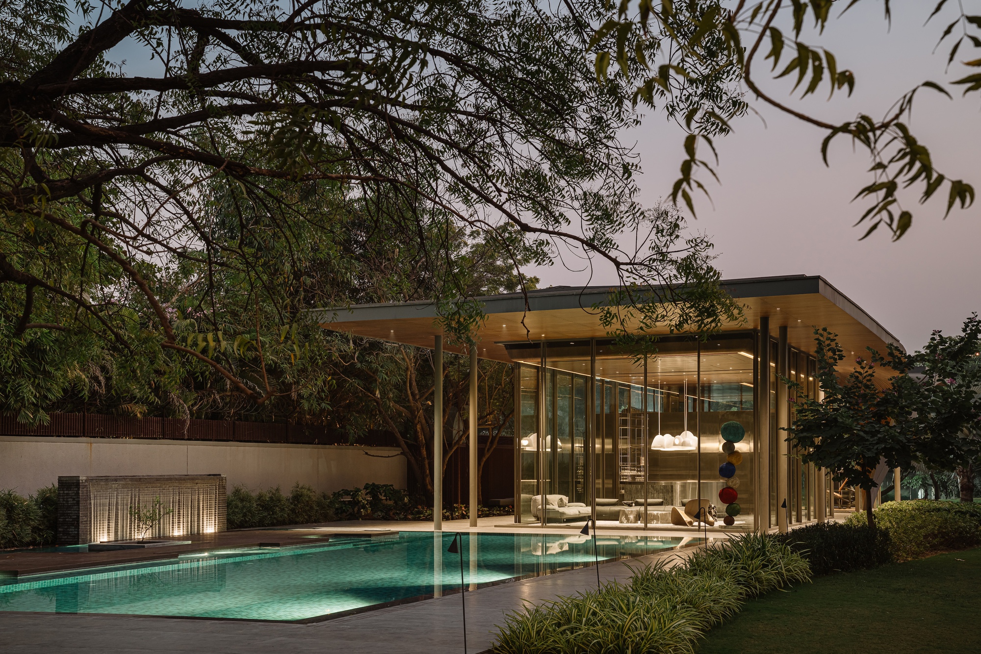 Evening view of glass pavilion beside pool framed by overhanging tree branches and garden planting