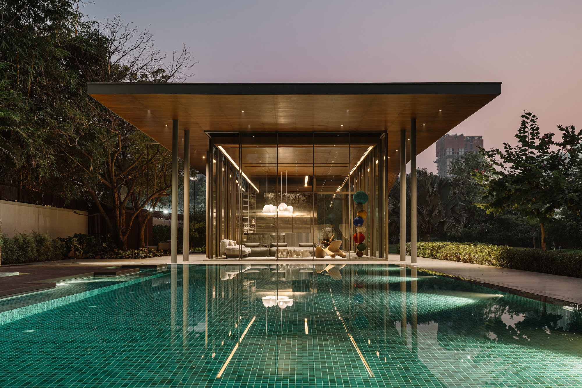 Glazed pavilion with flat overhang reflecting in swimming pool at dusk with interior lighting visible