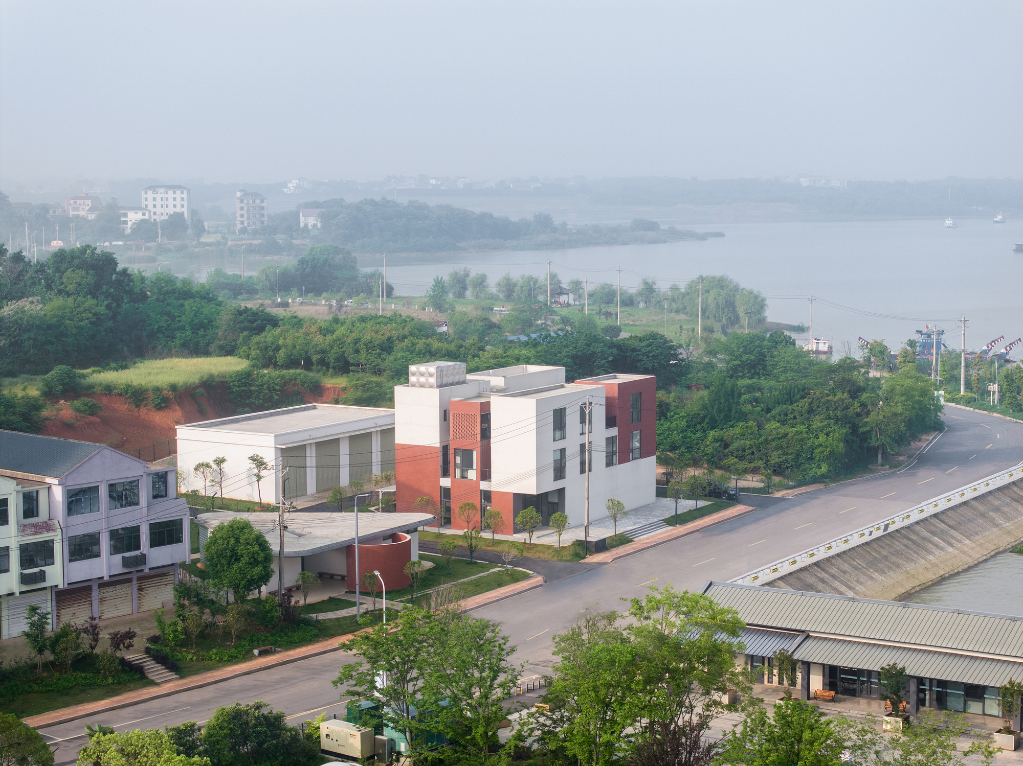 Aerial view of the multi-volume building with red brick and white plaster surrounded by waterfront greenery and mist