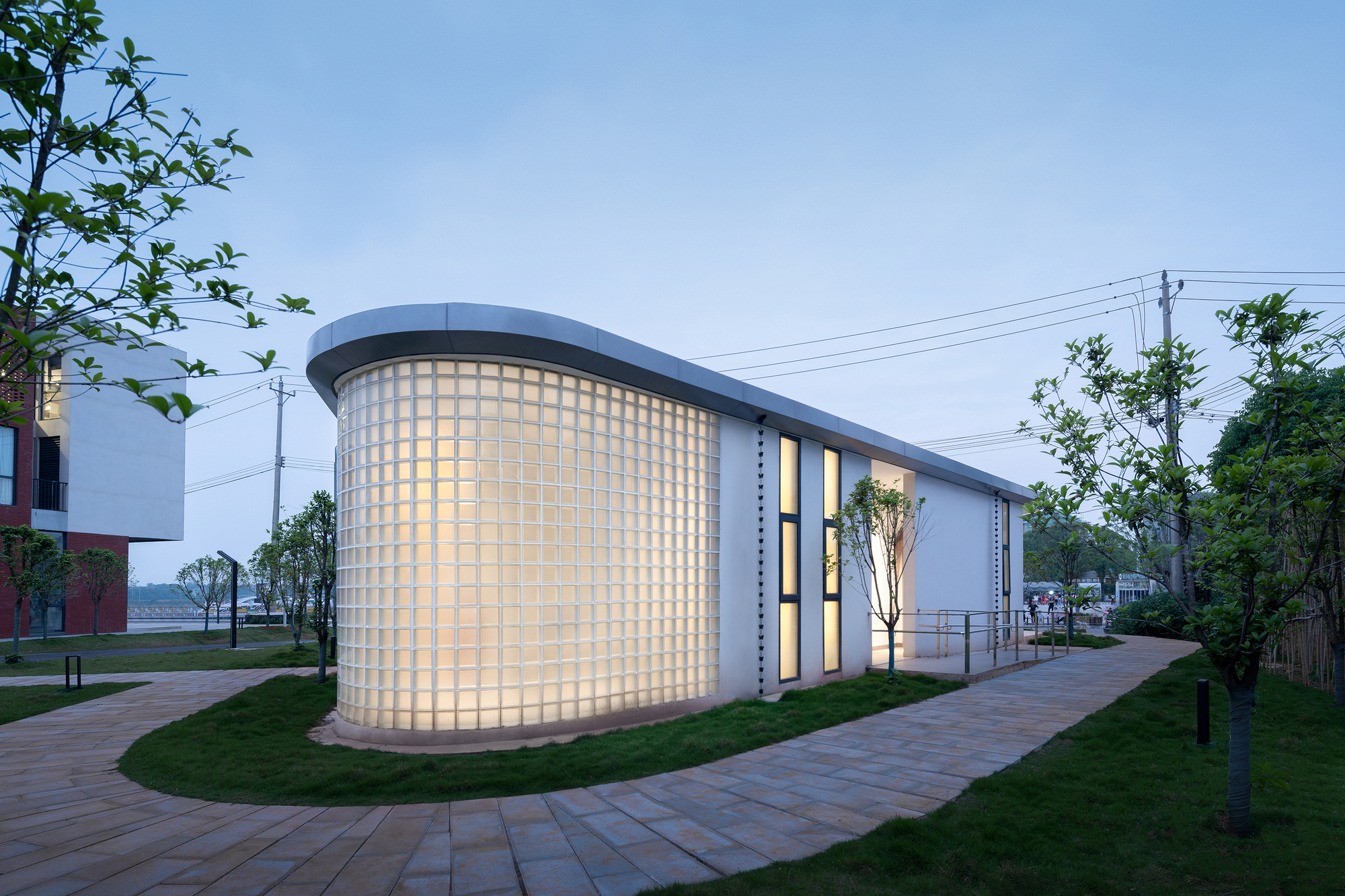 Curved translucent glass block wall illuminated at dusk along a paved pathway with trees