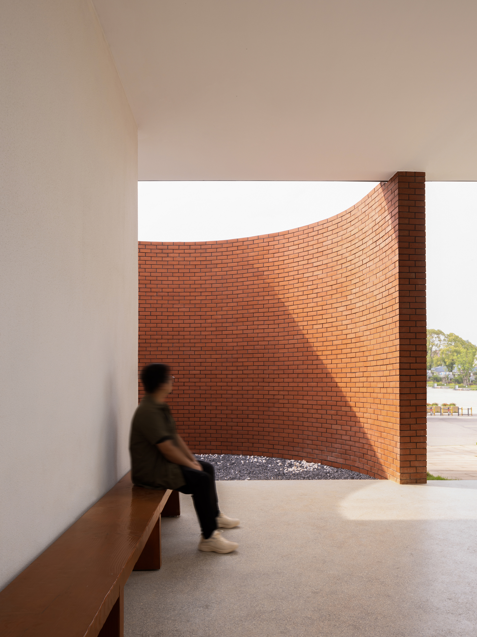 Interior view through white walls framing a curved red brick screen with person seated on timber bench