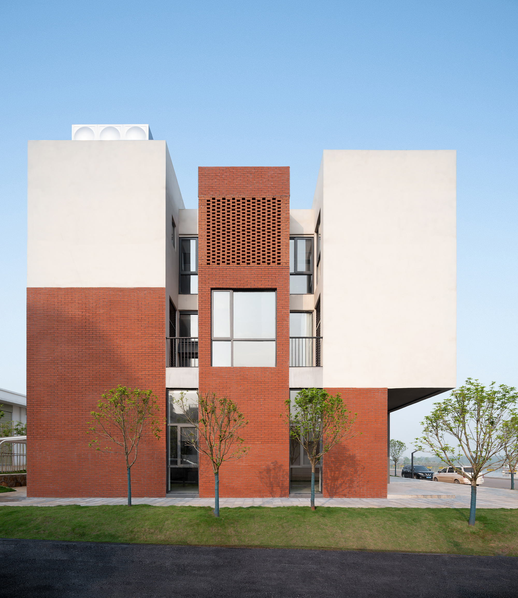 Front facade with red brick perforated screen centered between two white plaster volumes and young trees