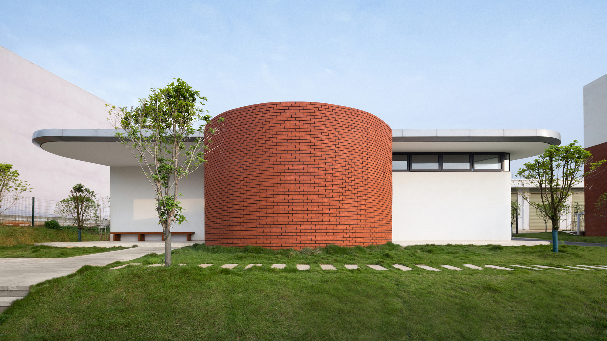 Cylindrical red brick volume with white stucco wings and flat roofs surrounded by young trees and grass pavers