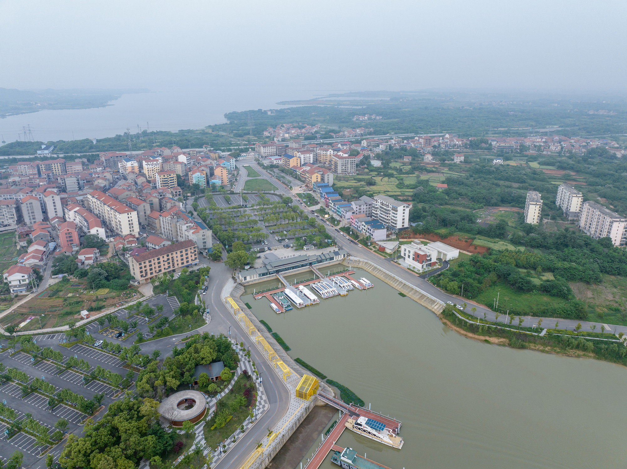Aerial view of a curved waterfront promenade with moored boats along a brown river and residential buildings beyond