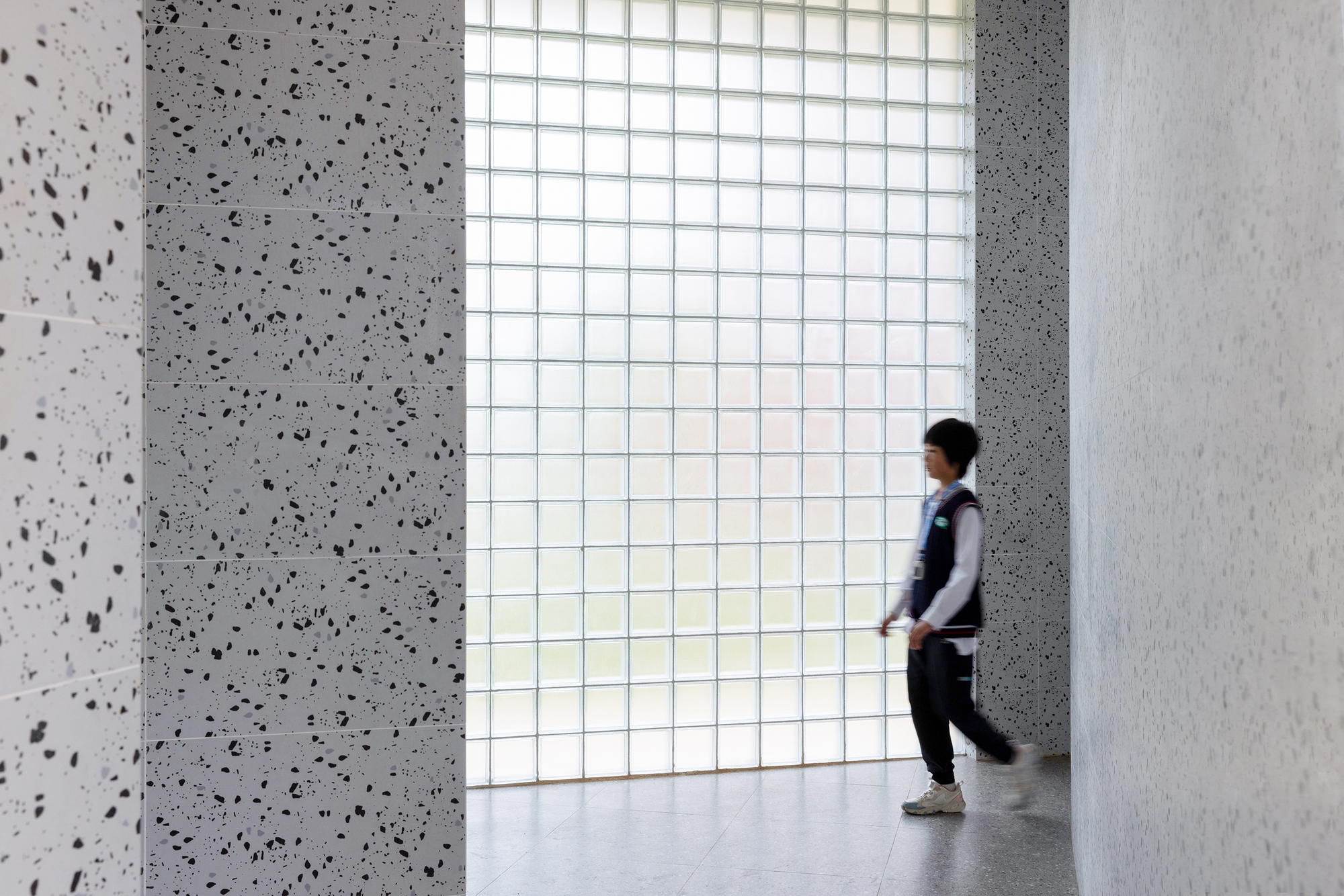 Interior corridor with terrazzo walls and translucent glass block wall as a person walks through diffused daylight