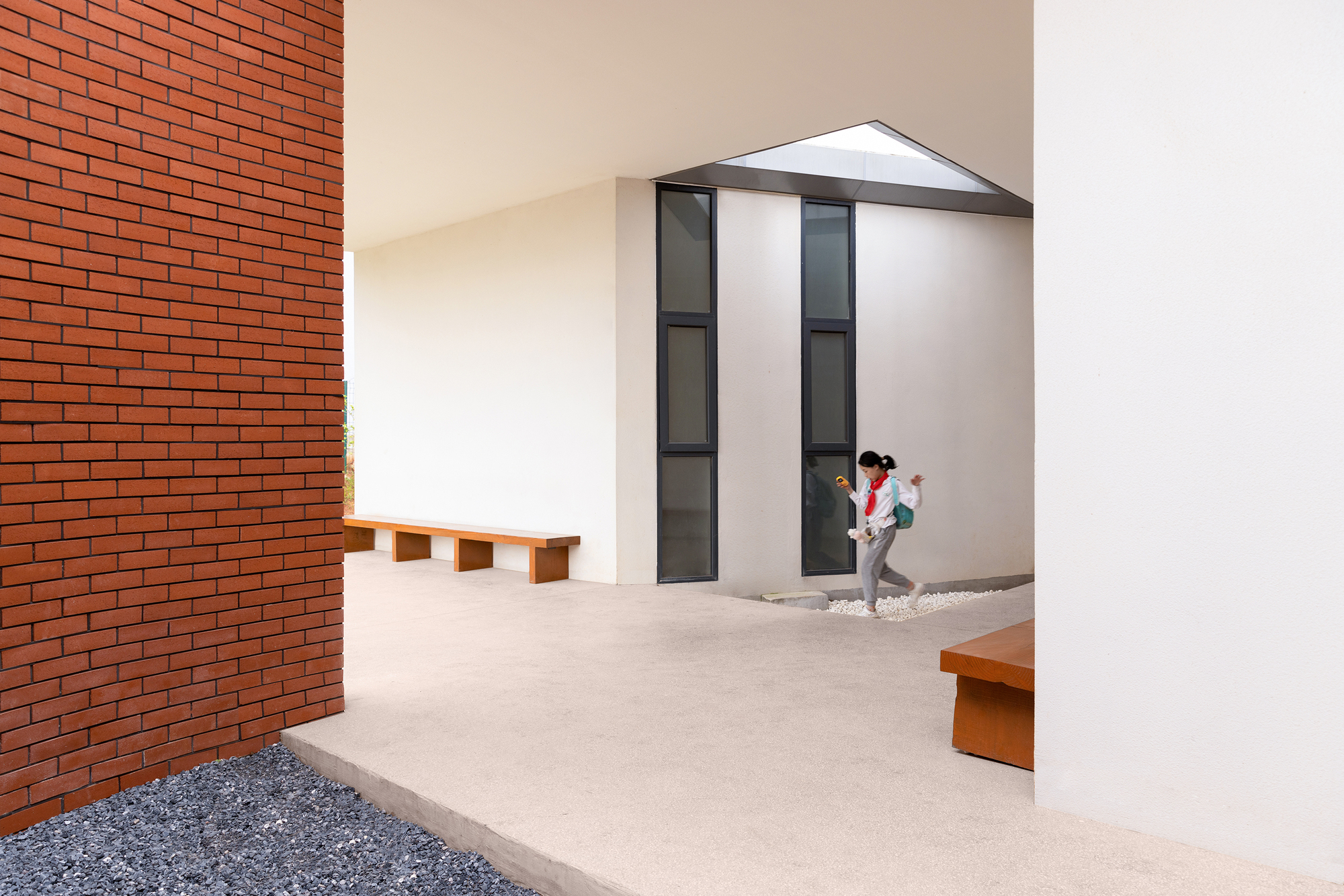 Courtyard with red brick wall and timber benches framing a white stucco volume with vertical glazing and gravel