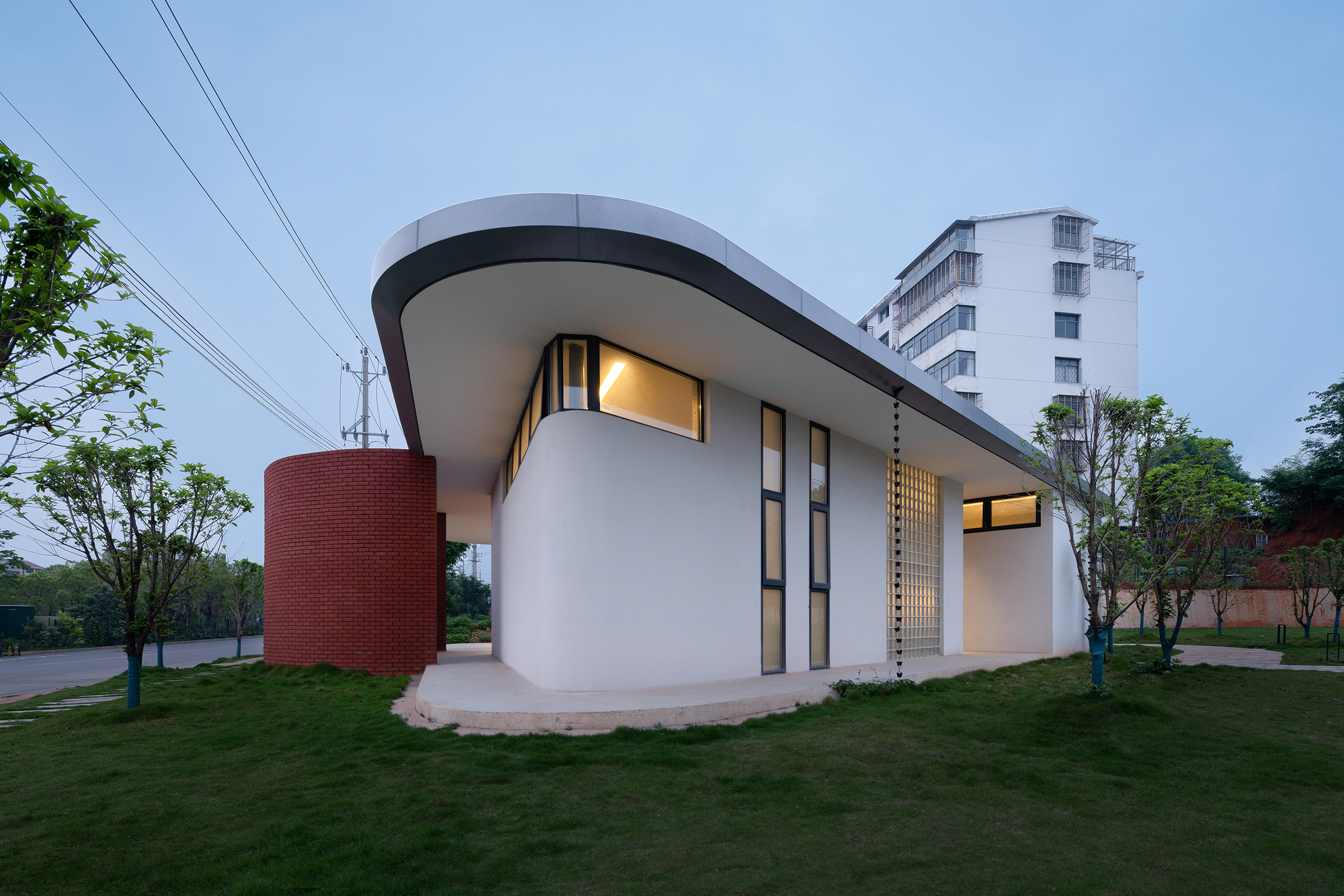 Corner view of white stucco volume with red brick cylinder and cantilevered roof at dusk with interior lights glowing