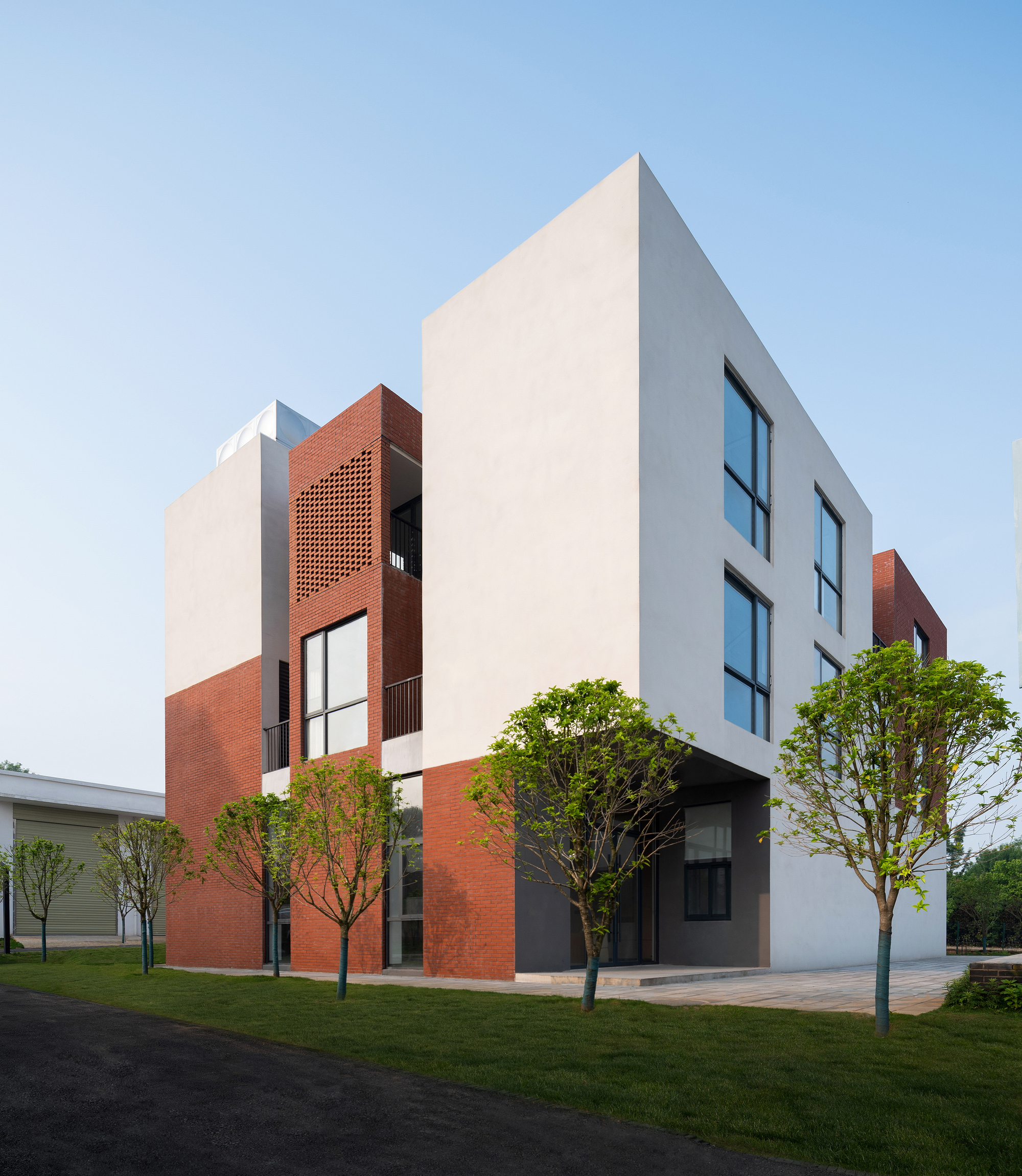 Street facade combining white stucco volumes with perforated red brick screens and cantilevered upper floor framed by young trees