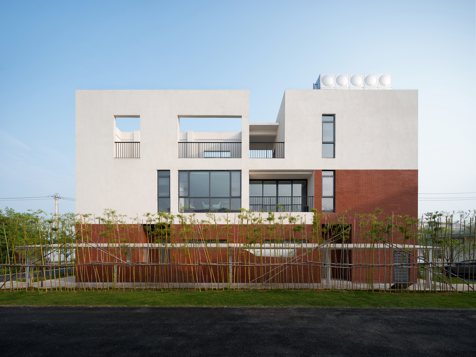 Street view of the two-story facade with white stucco and red brick base behind a young bamboo planting