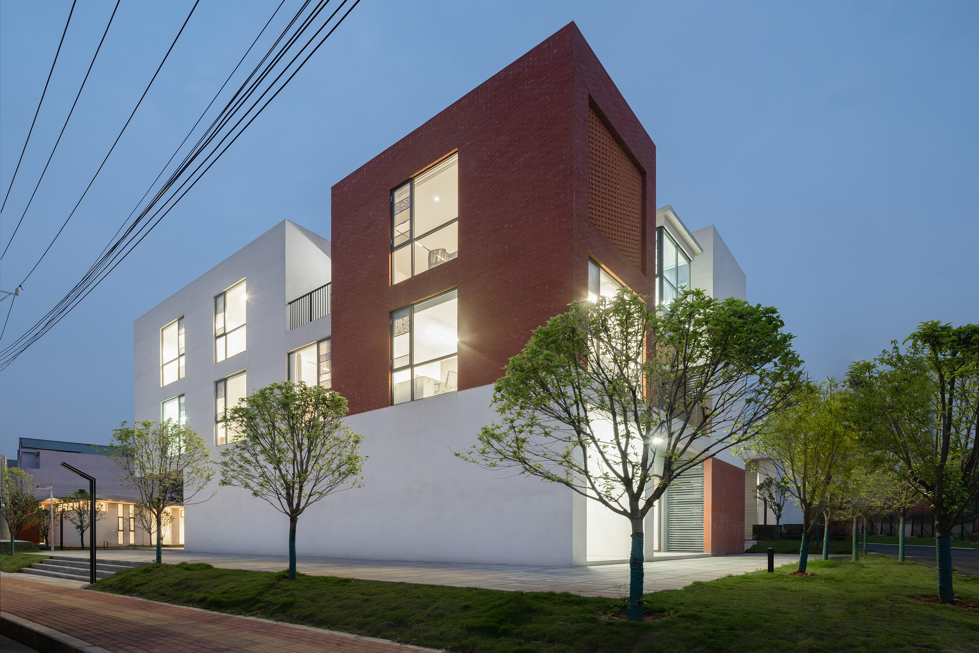Corner view at twilight showing the red brick volumes and white stucco with interior lights glowing