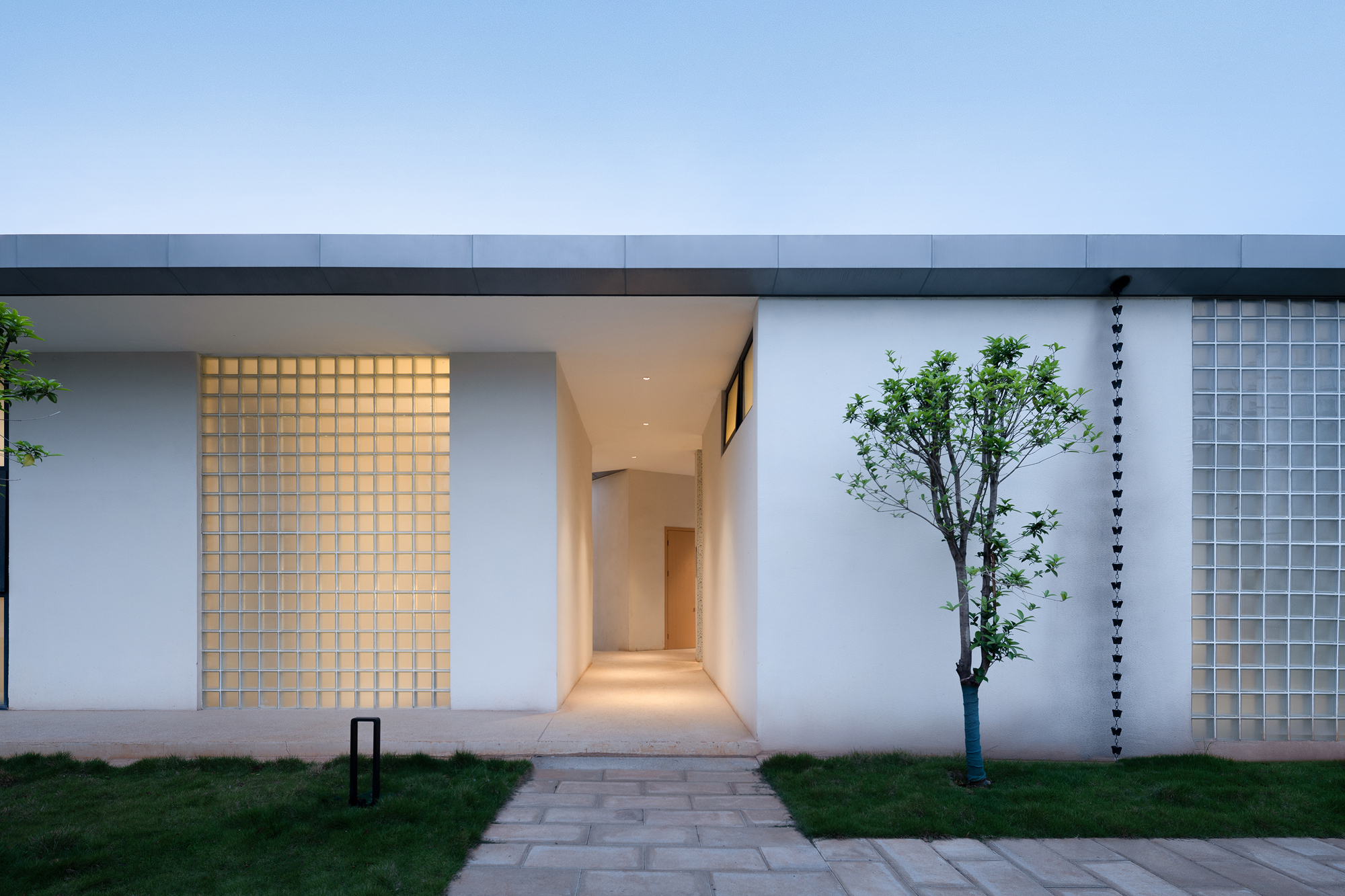 Entry facade at dusk showing glass block screen and illuminated corridor with a courtyard tree