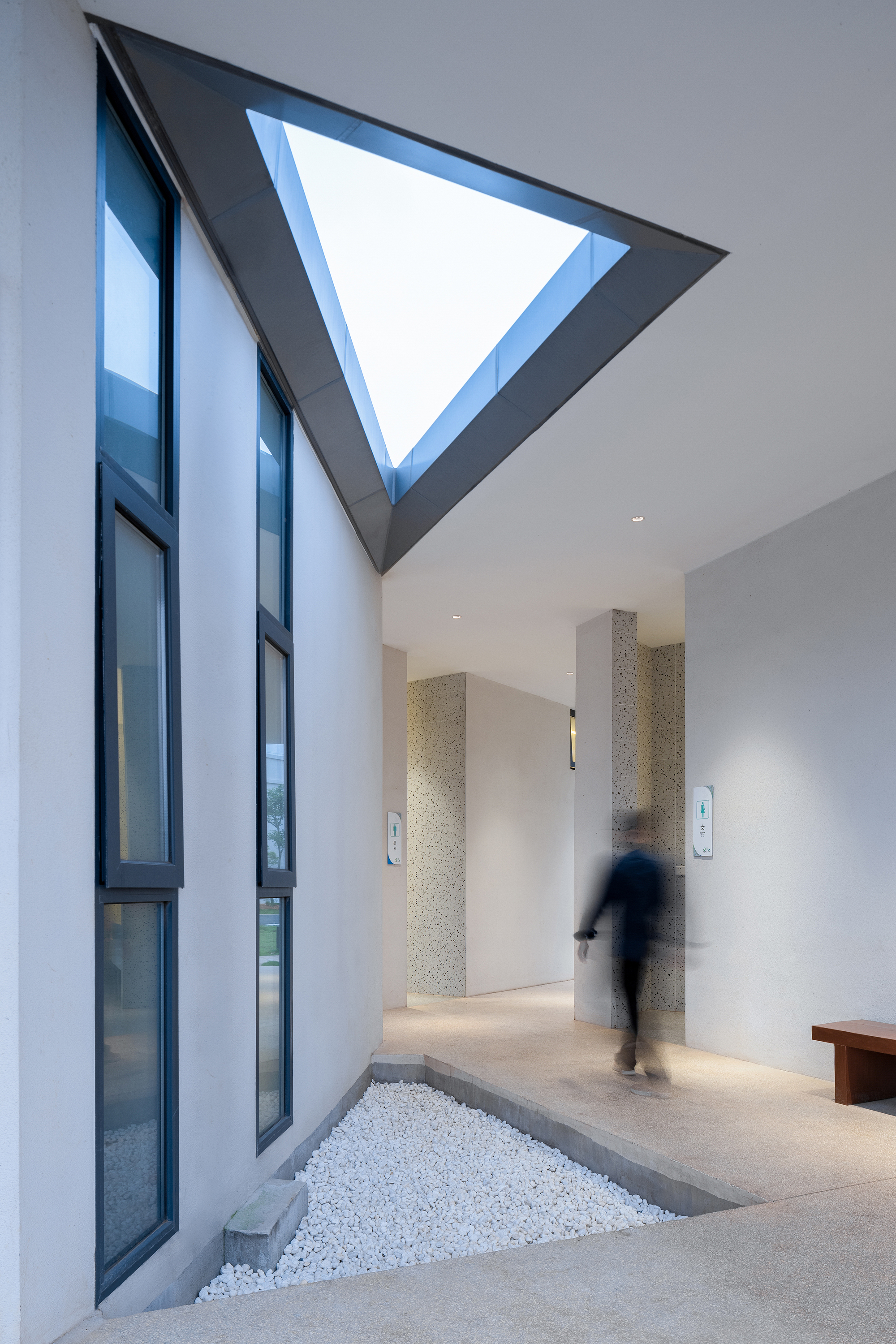 Double-height corridor with triangular skylight above and a figure walking past white pebble bed