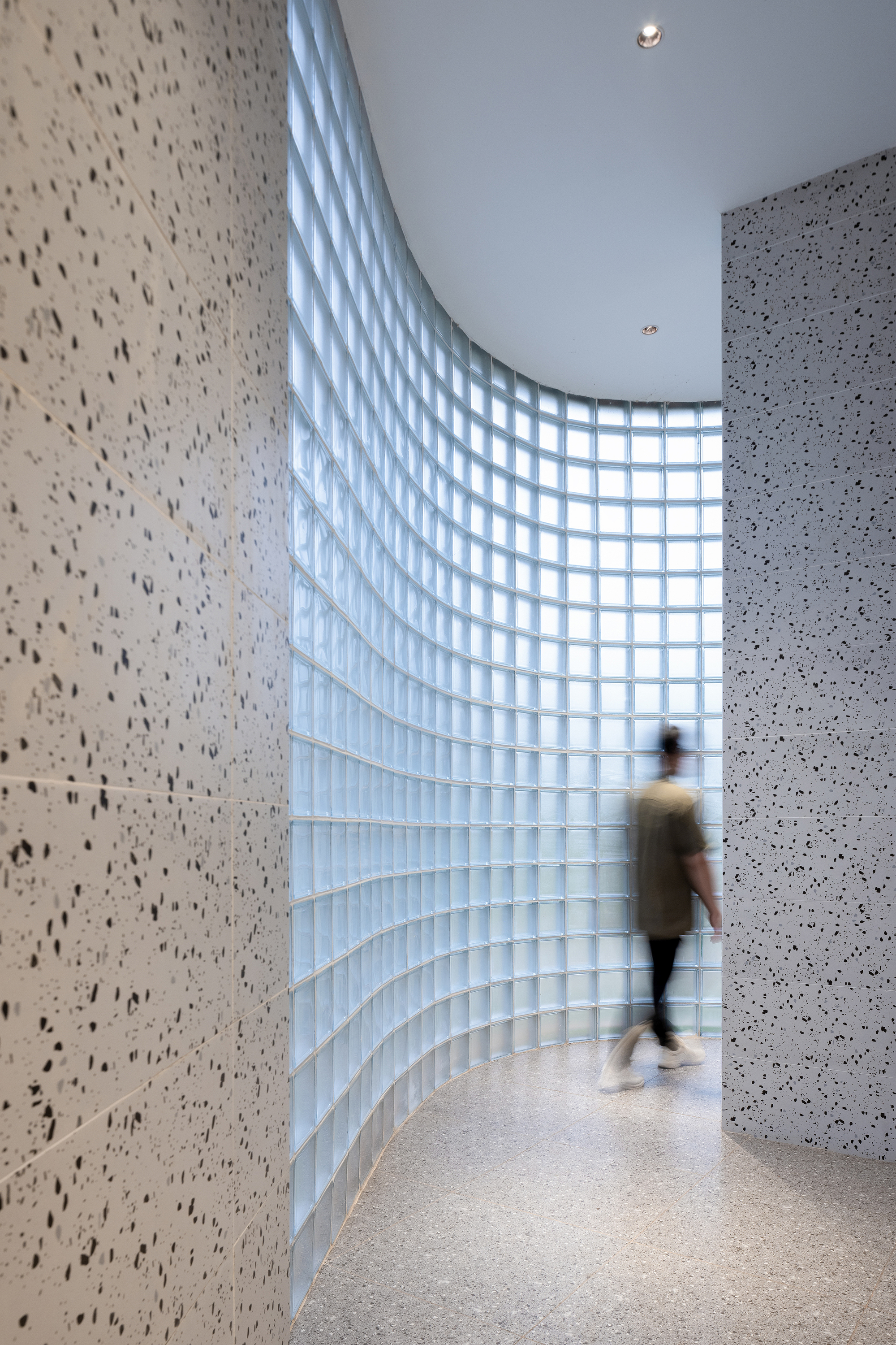 Curved glass block wall along a corridor with terrazzo panels and a visitor in motion