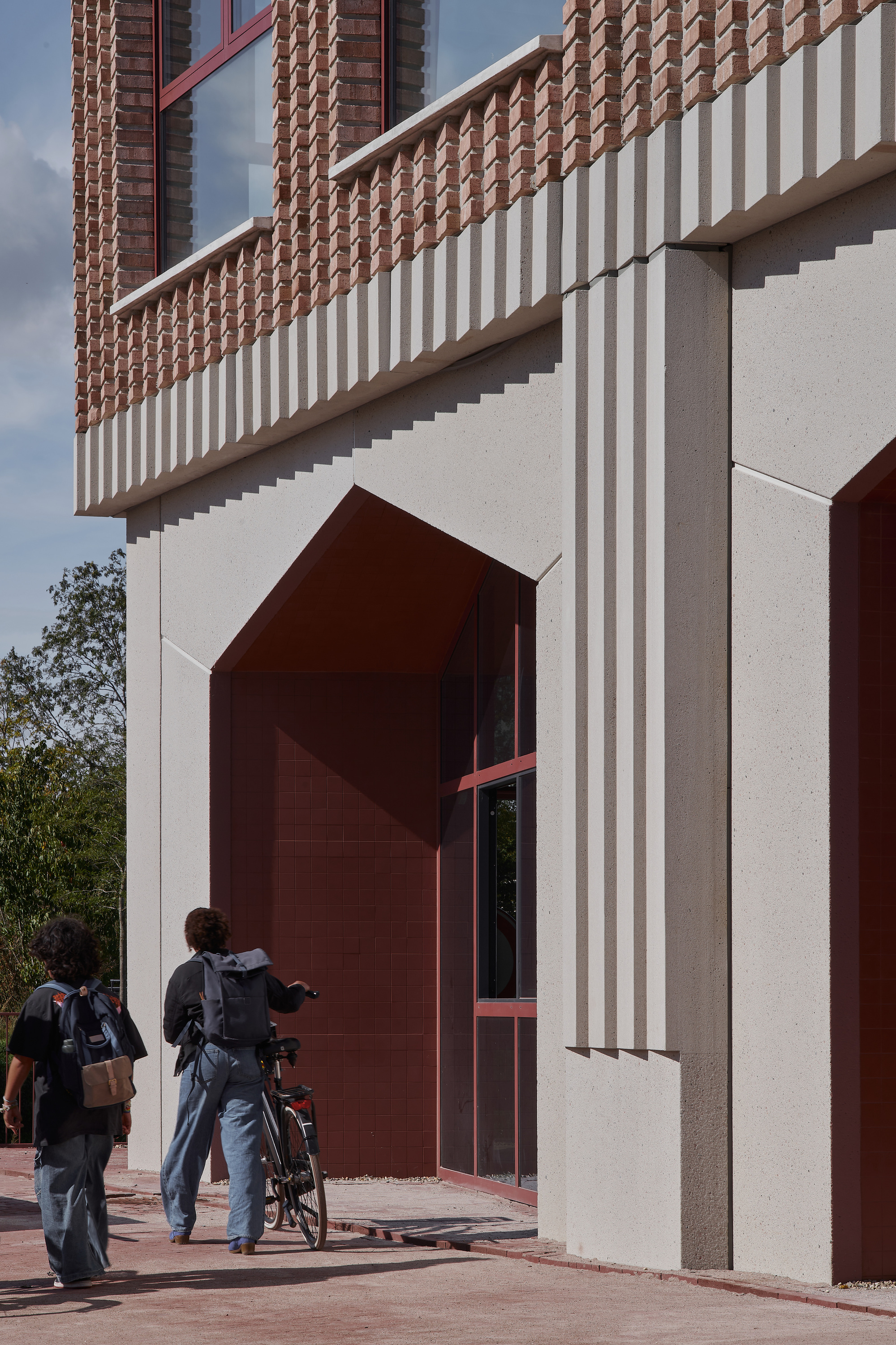 Close-up of arched maroon entrance portal with vertical fluted precast cladding and people approaching with bicycles