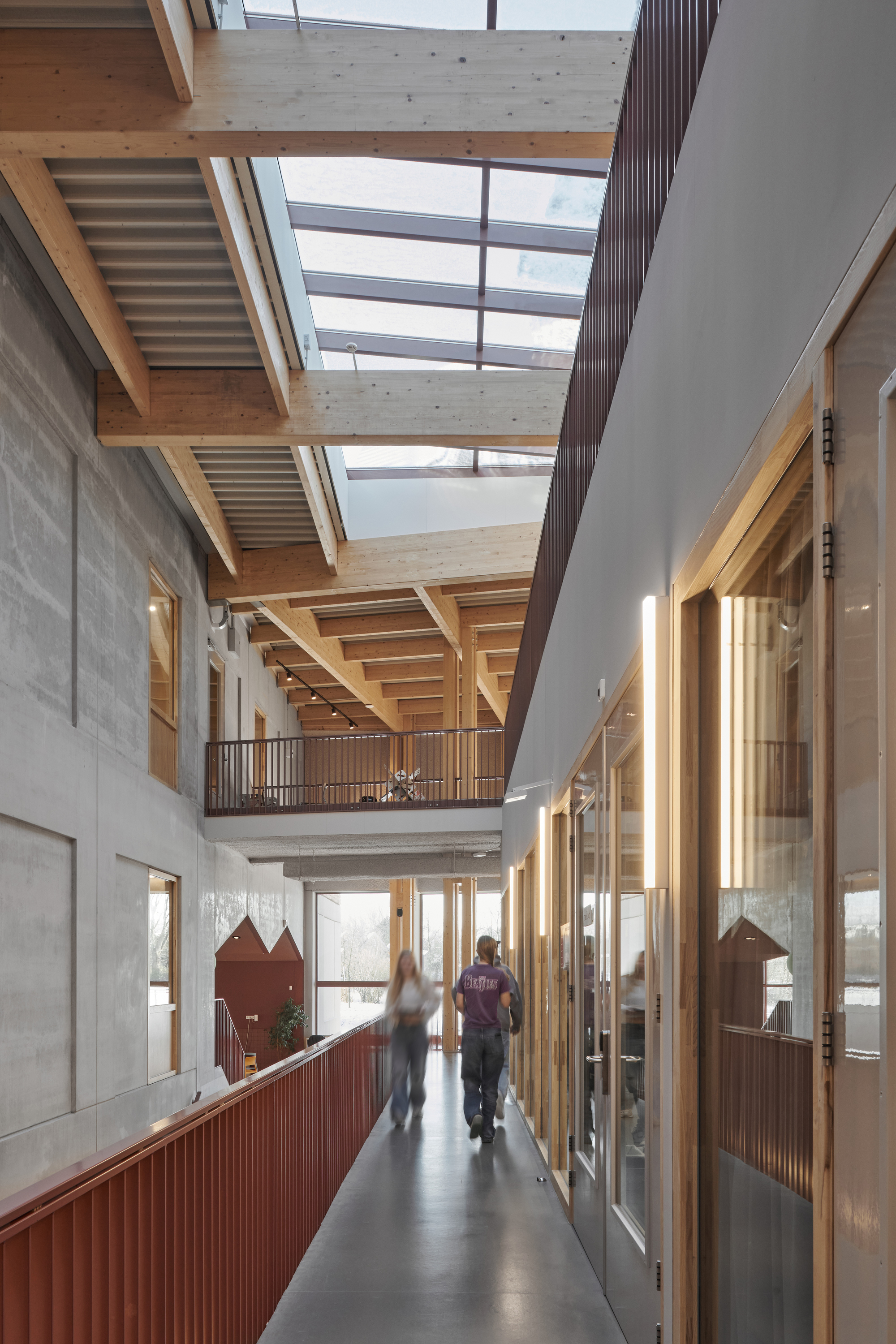 Upper corridor with timber beam ceiling, skylights, concrete walls, and maroon-paneled balustrades overlooking the atrium