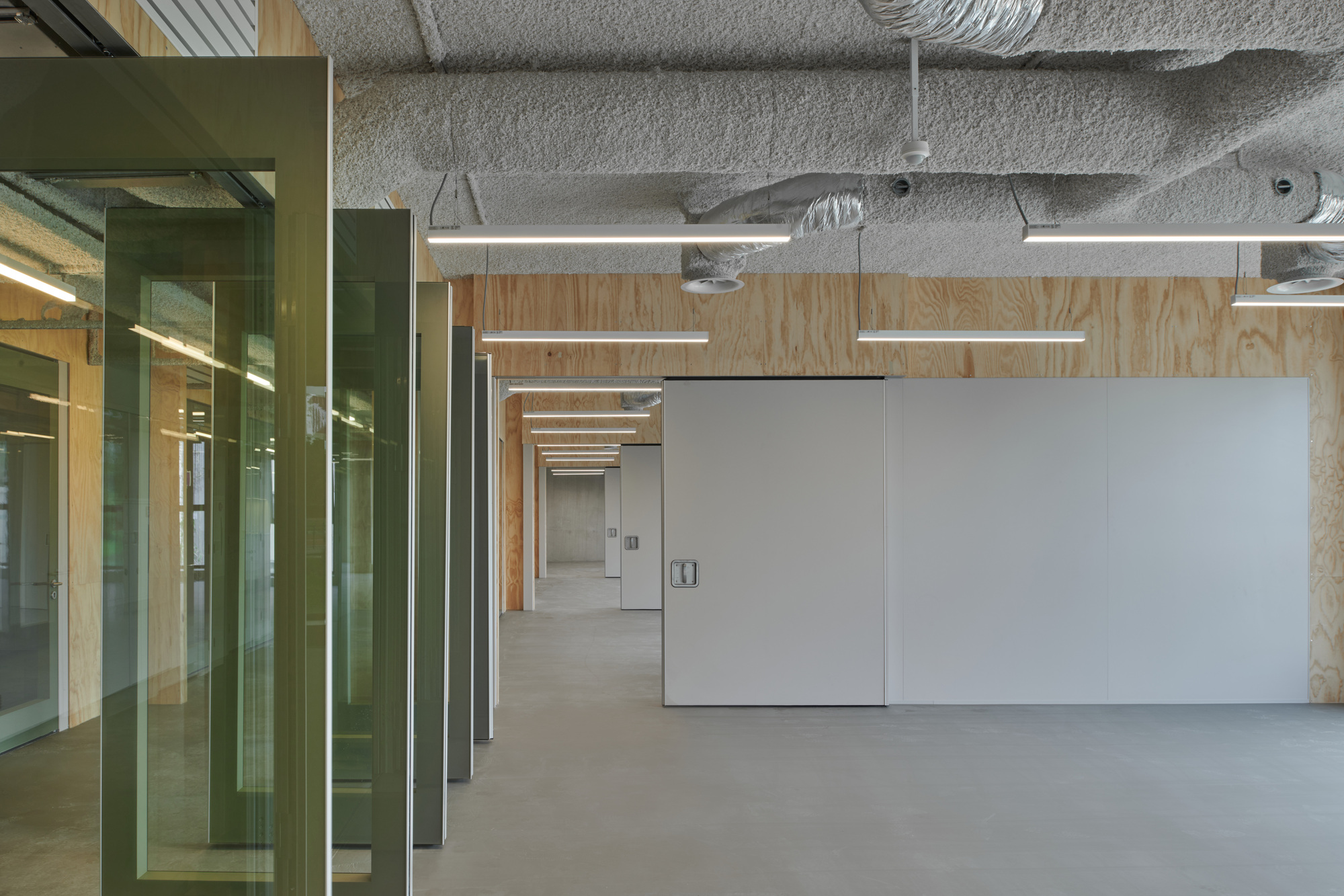 Open office space with exposed ductwork ceiling and white partition walls flanked by green glass enclosures