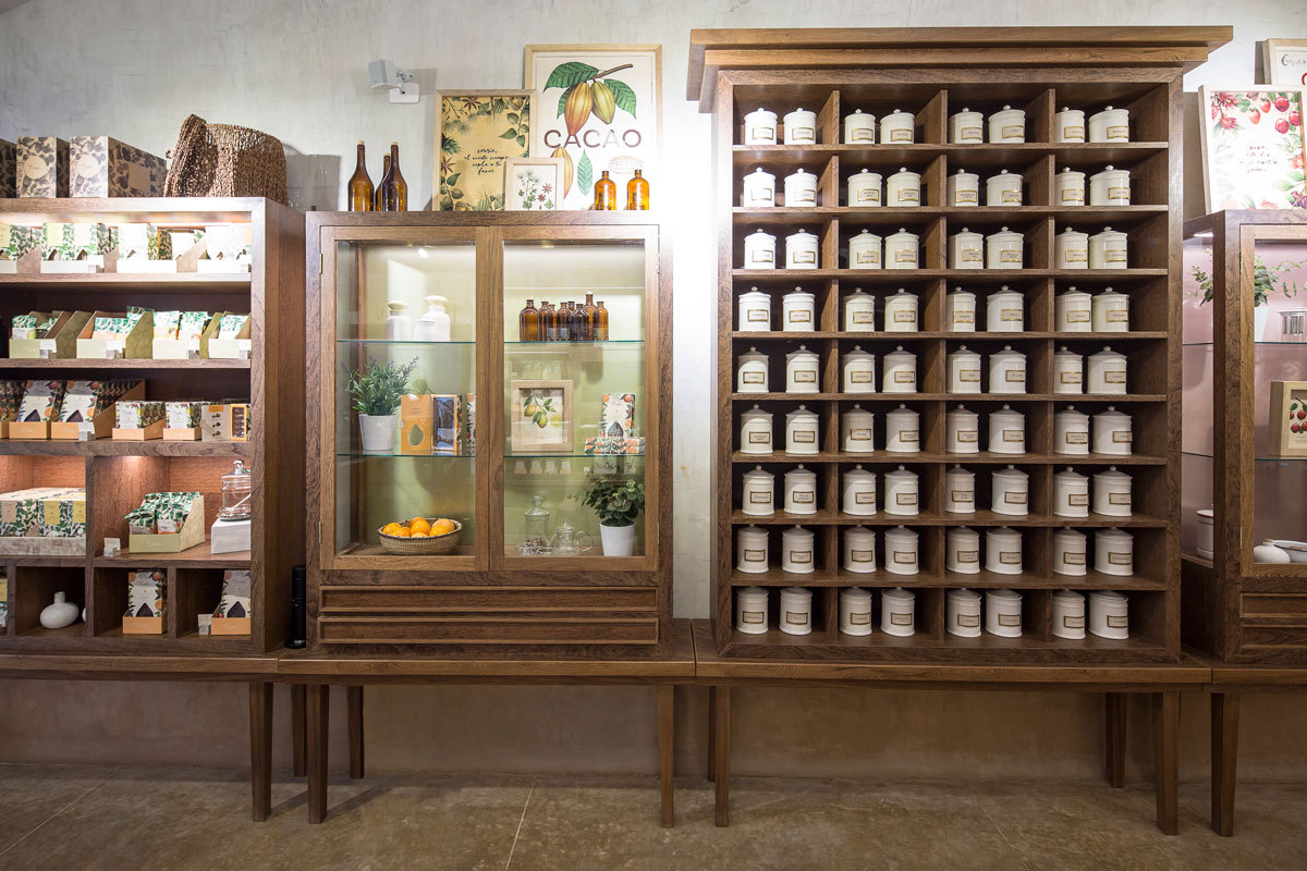 Wall of dark timber display cabinets holding rows of white lidded canisters beside a glass vitrine with potted plants and fruit