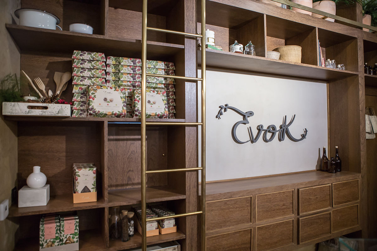 Timber shelving wall with a brass ladder, boxed goods, and a pale panel carrying a script wordmark above drawers