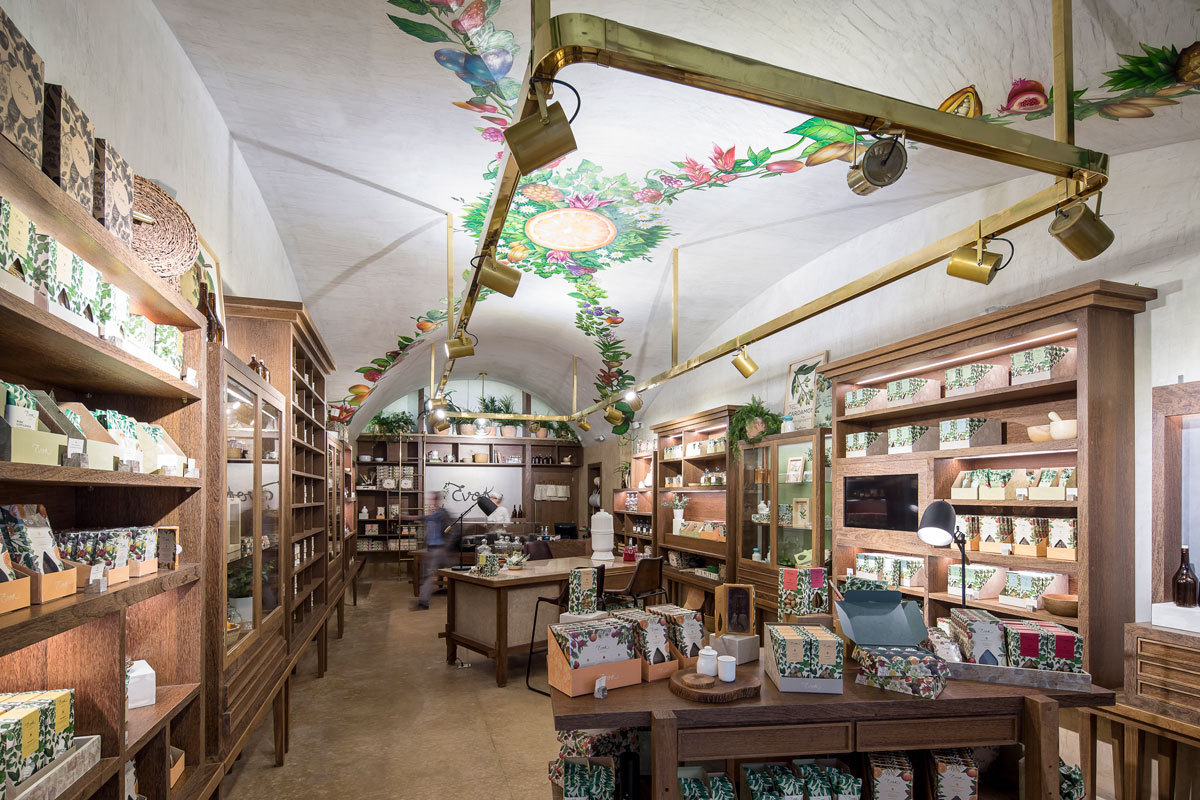Vaulted shop interior with a painted fruit and flower ceiling, brass track lighting, and a passerby blurred near the central table
