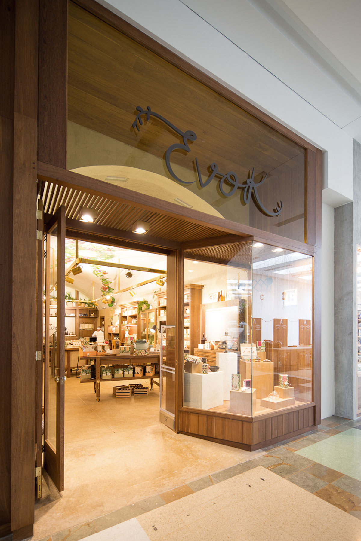 Shopfront entrance with a timber frame, glass display window, and hanging script sign, seen from the mall floor in warm light
