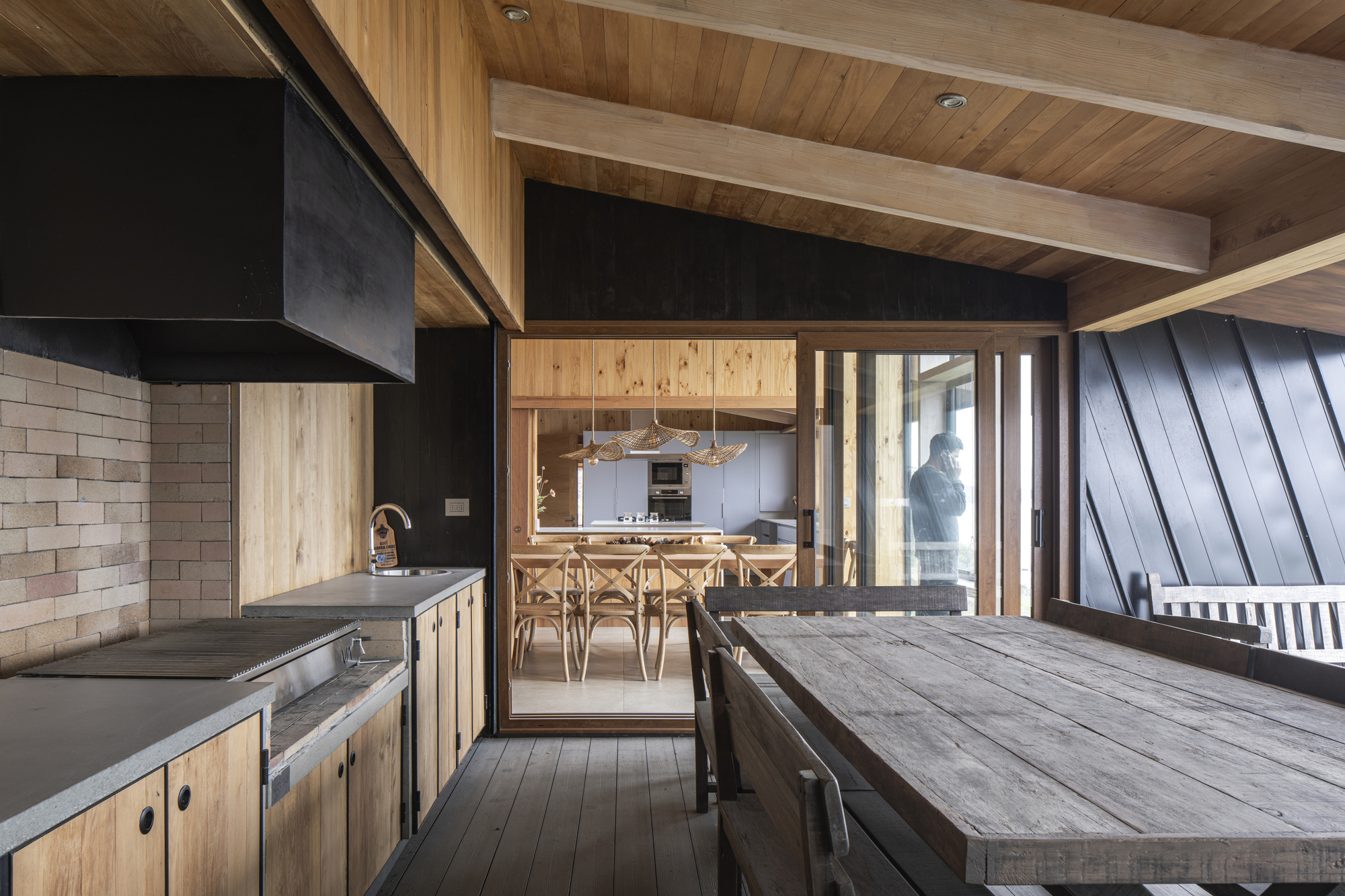 Interior dining area with timber ceiling beams and recycled wood table looking through to kitchen beneath dark range hood
