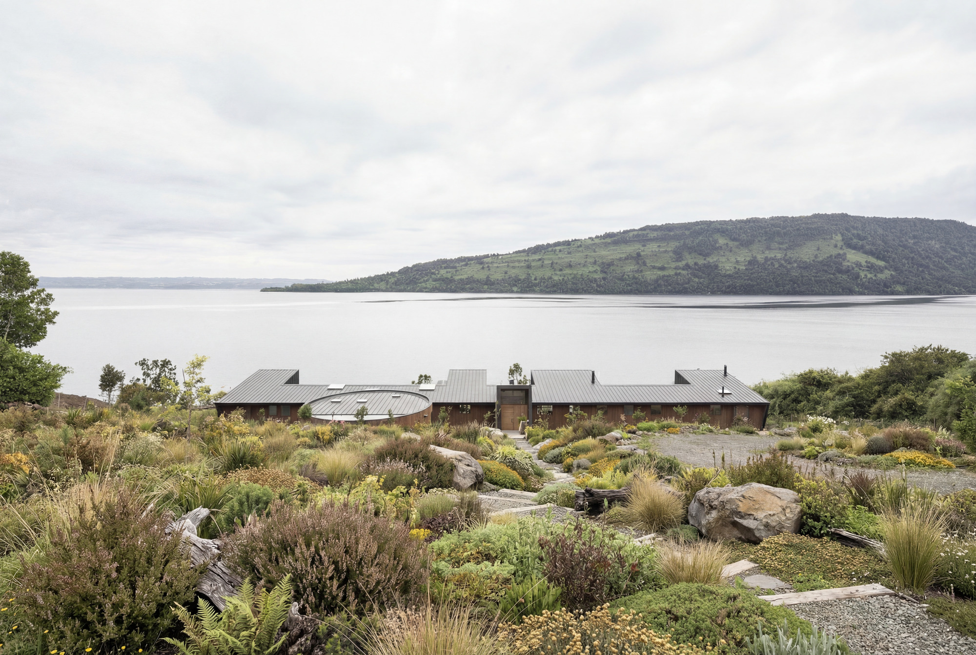 Low-profile metal-roofed volumes nestled into a rocky hillside with native planting overlooking a calm lake and forested peninsula