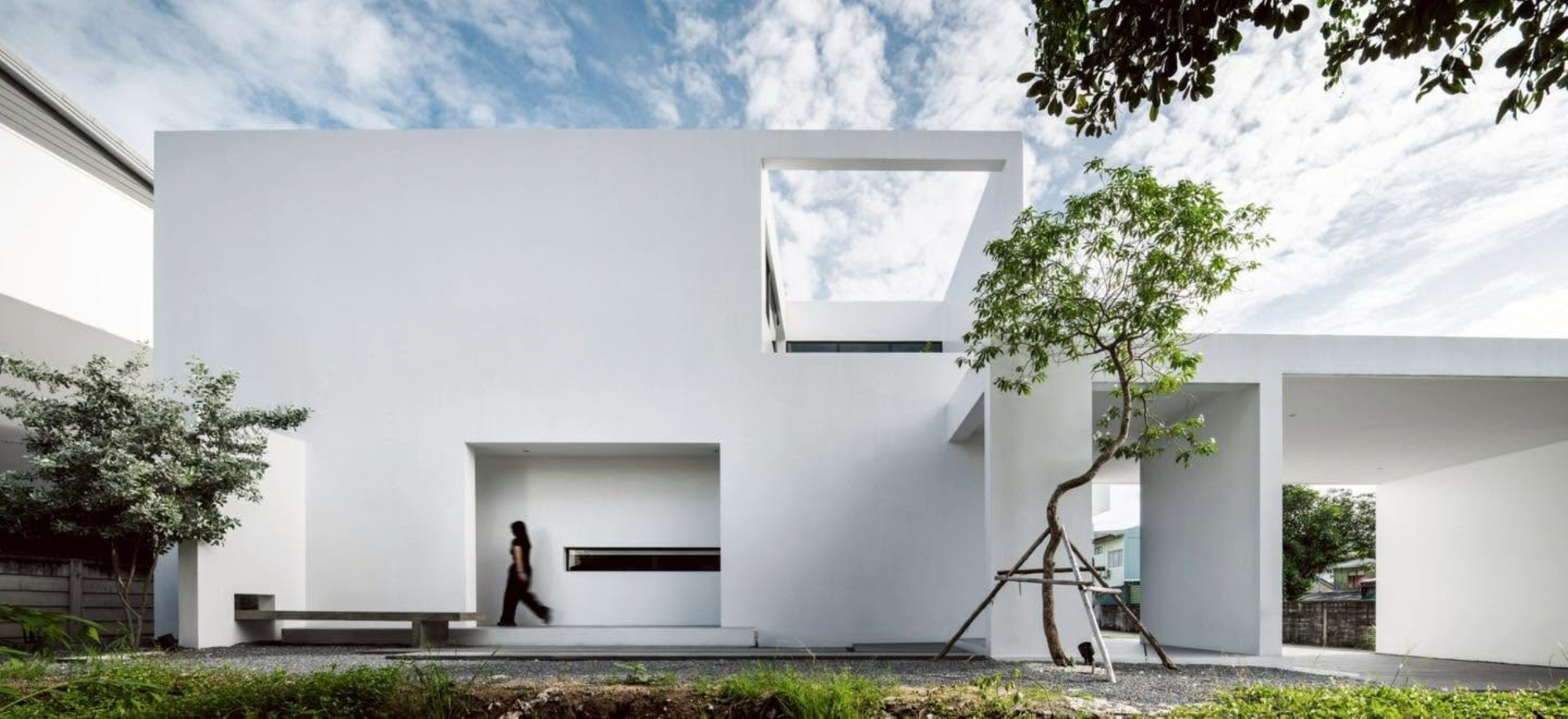 White cubic volumes with framed openings and a planted tree beside a gravel courtyard
