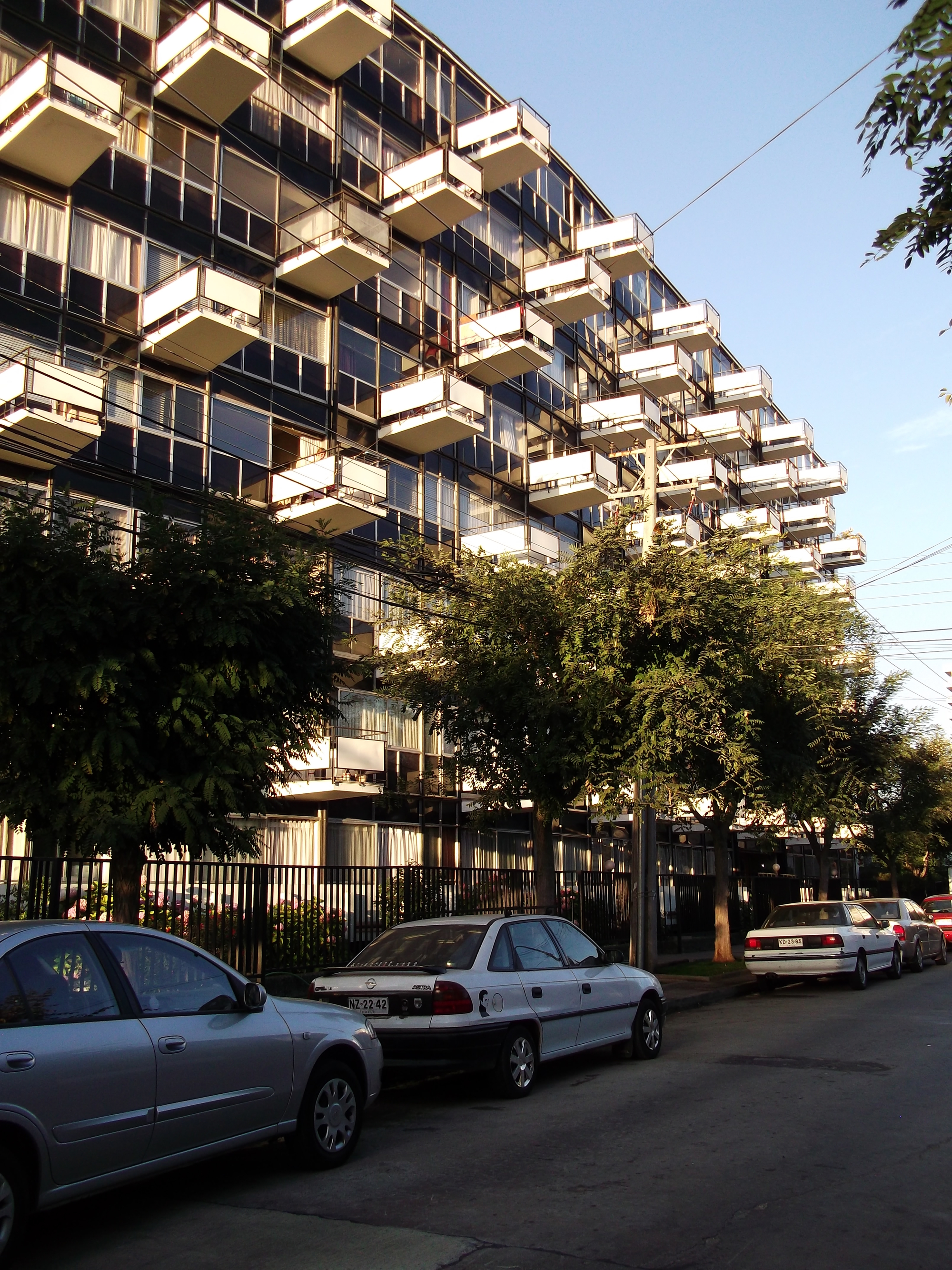 Street view of the residential tower with projecting balconies behind parked cars and mature trees