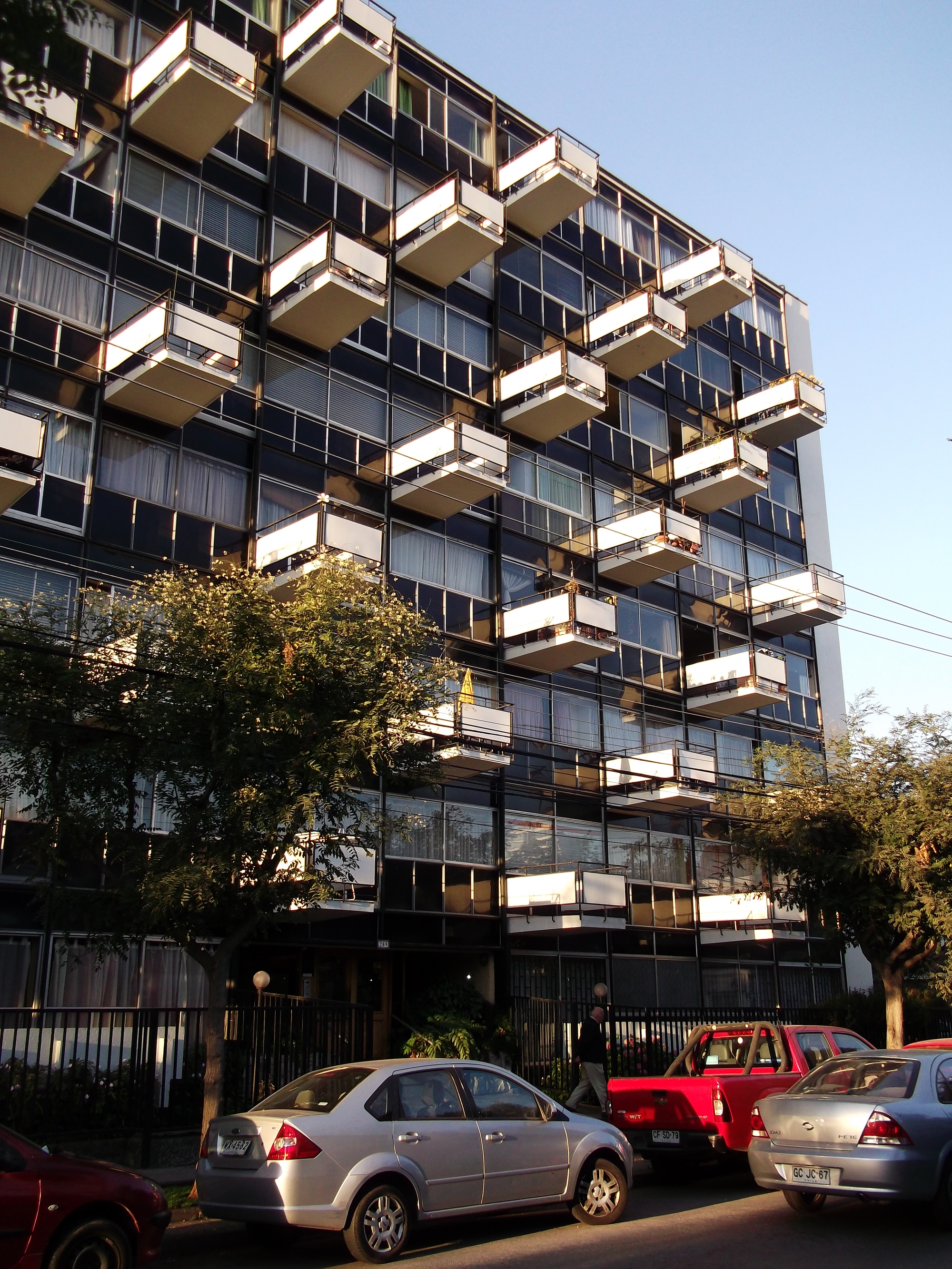 Street view of the residential tower facade with staggered concrete balconies and dark glazing framed by trees