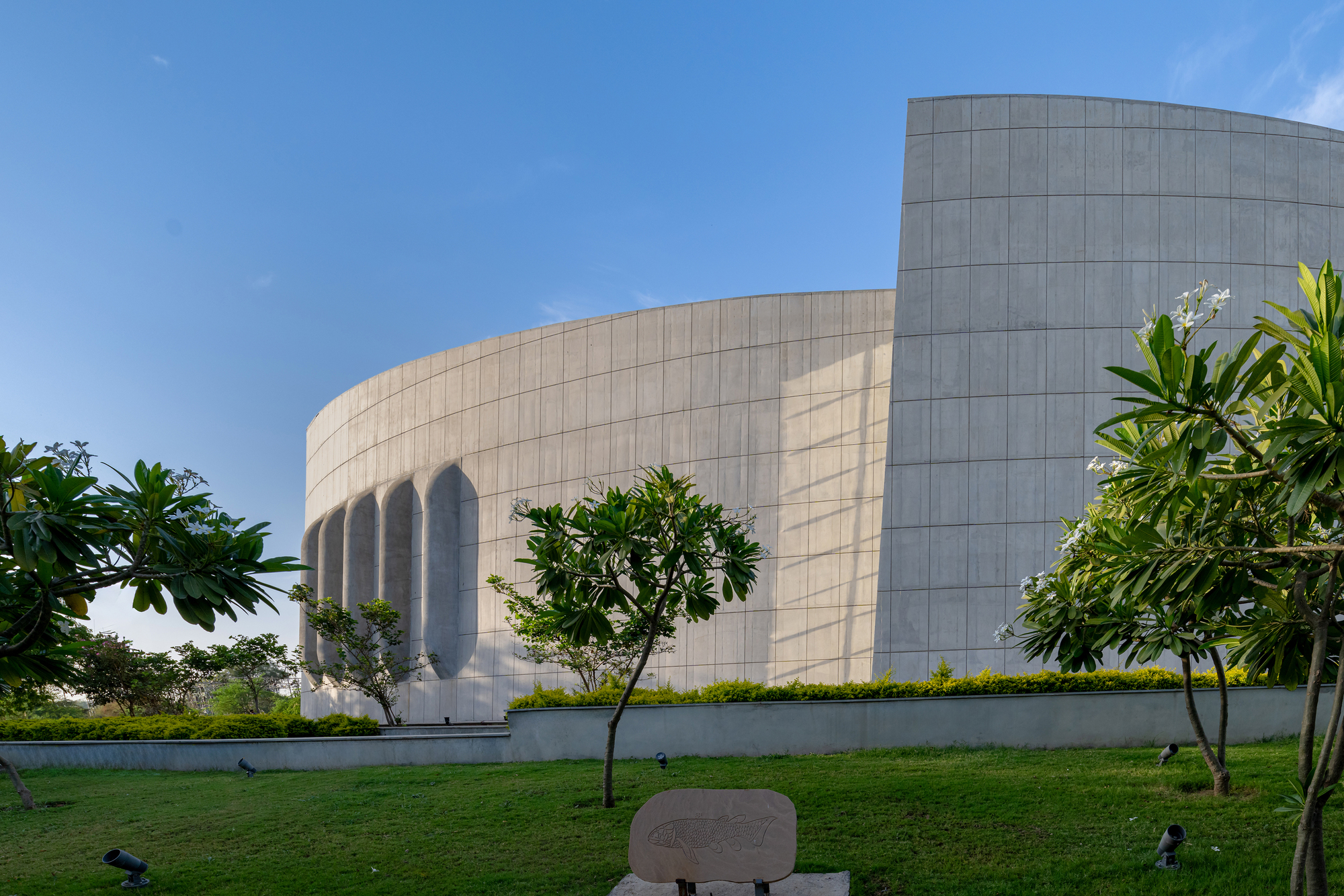 Ground-level view of the paneled stone facade with recessed arched openings behind frangipani trees and lawn