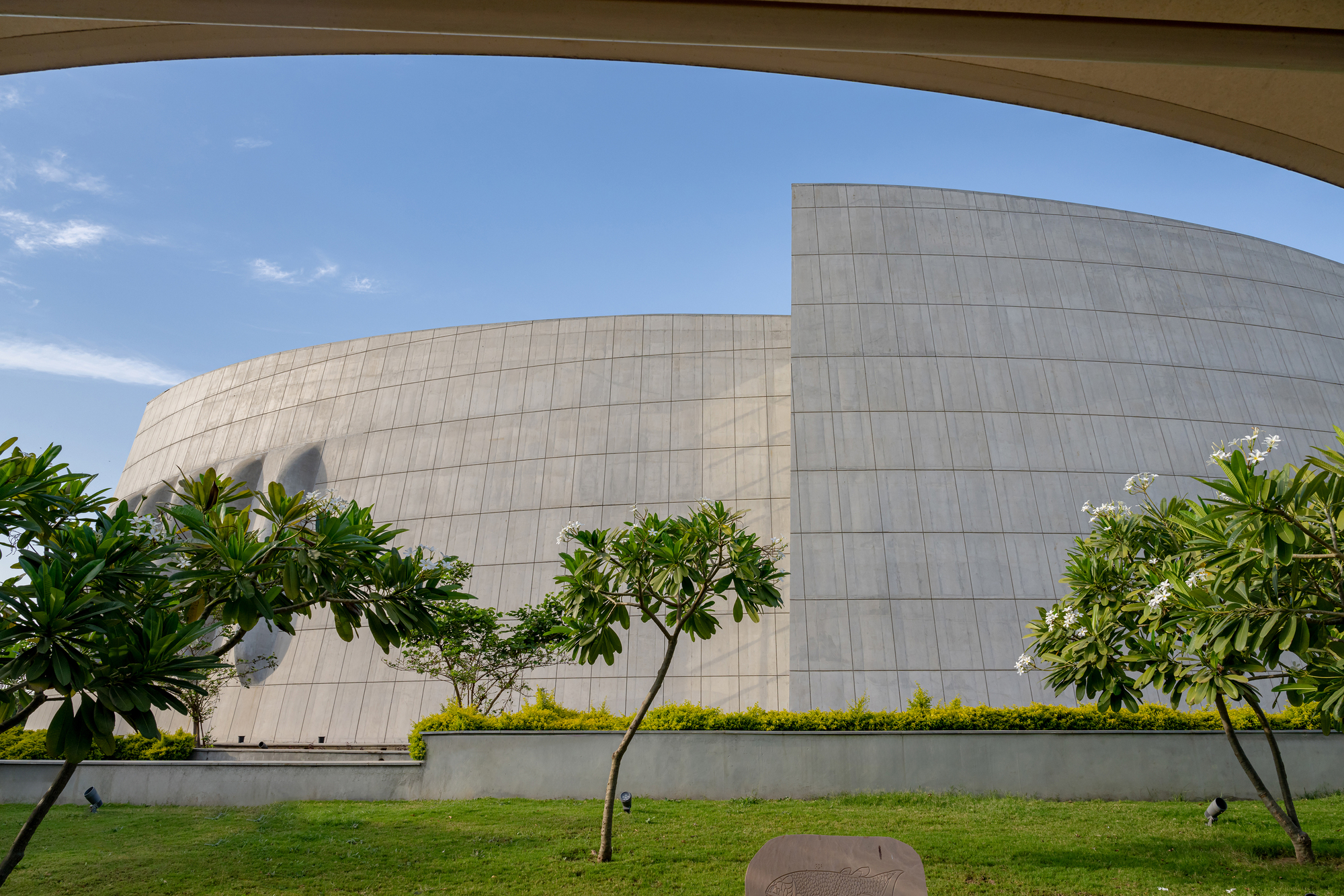 Stone-clad curved facade with planted young trees viewed through an overhead canopy