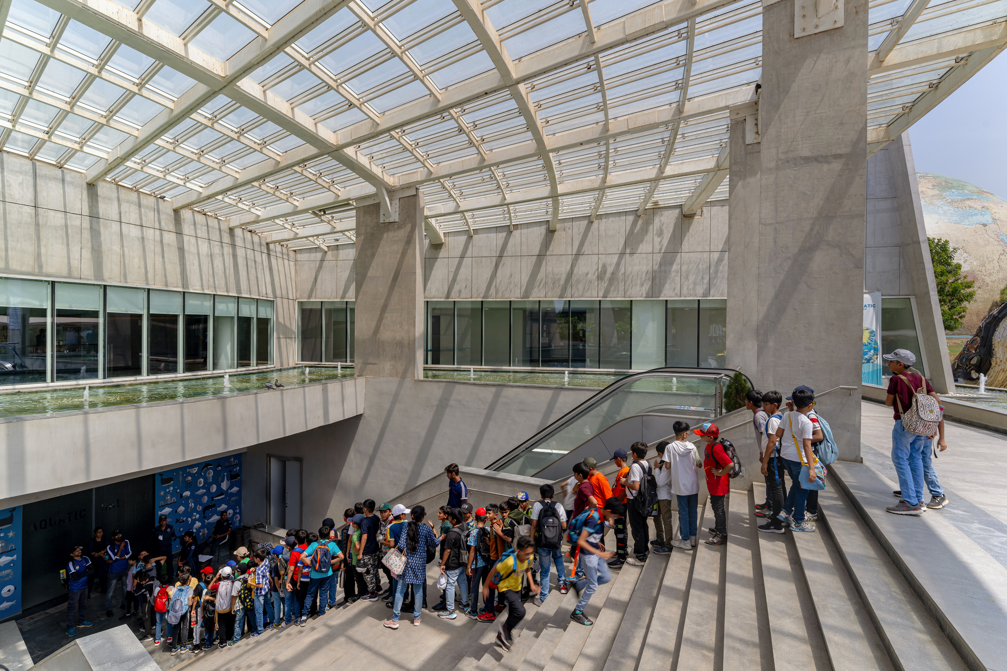 Double-height atrium with gridded glass canopy and concrete columns casting shadows over visitors queuing on an escalator
