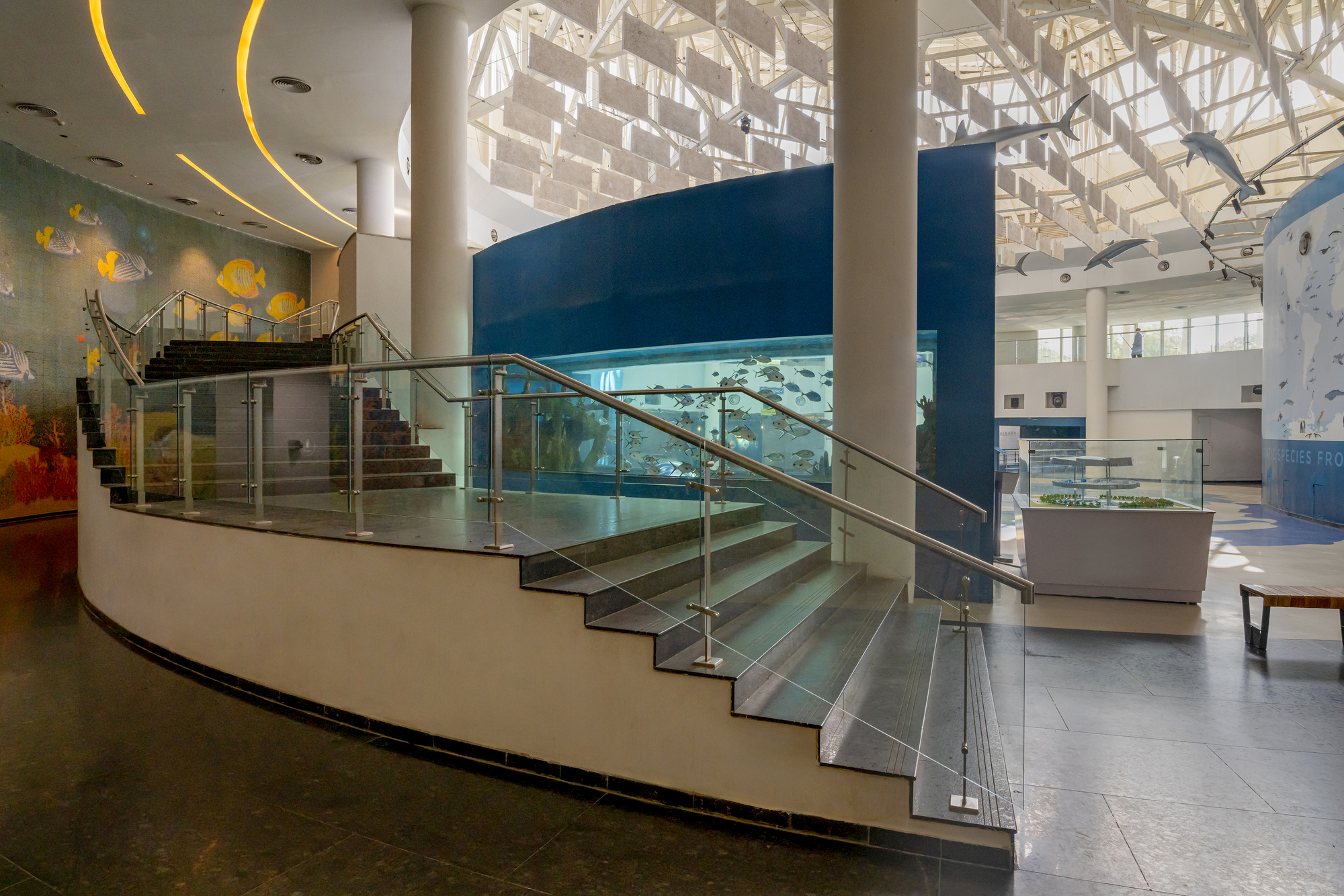 Curved staircase with glass balustrades and metal handrails beneath a geometric glass roof structure and yellow ceiling details