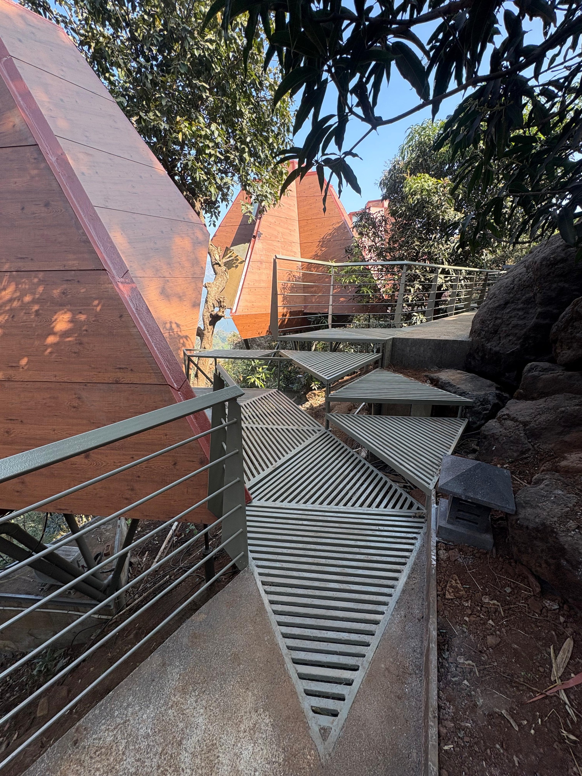 Metal walkway with horizontal railings weaving between pyramidal timber structures and boulders among trees