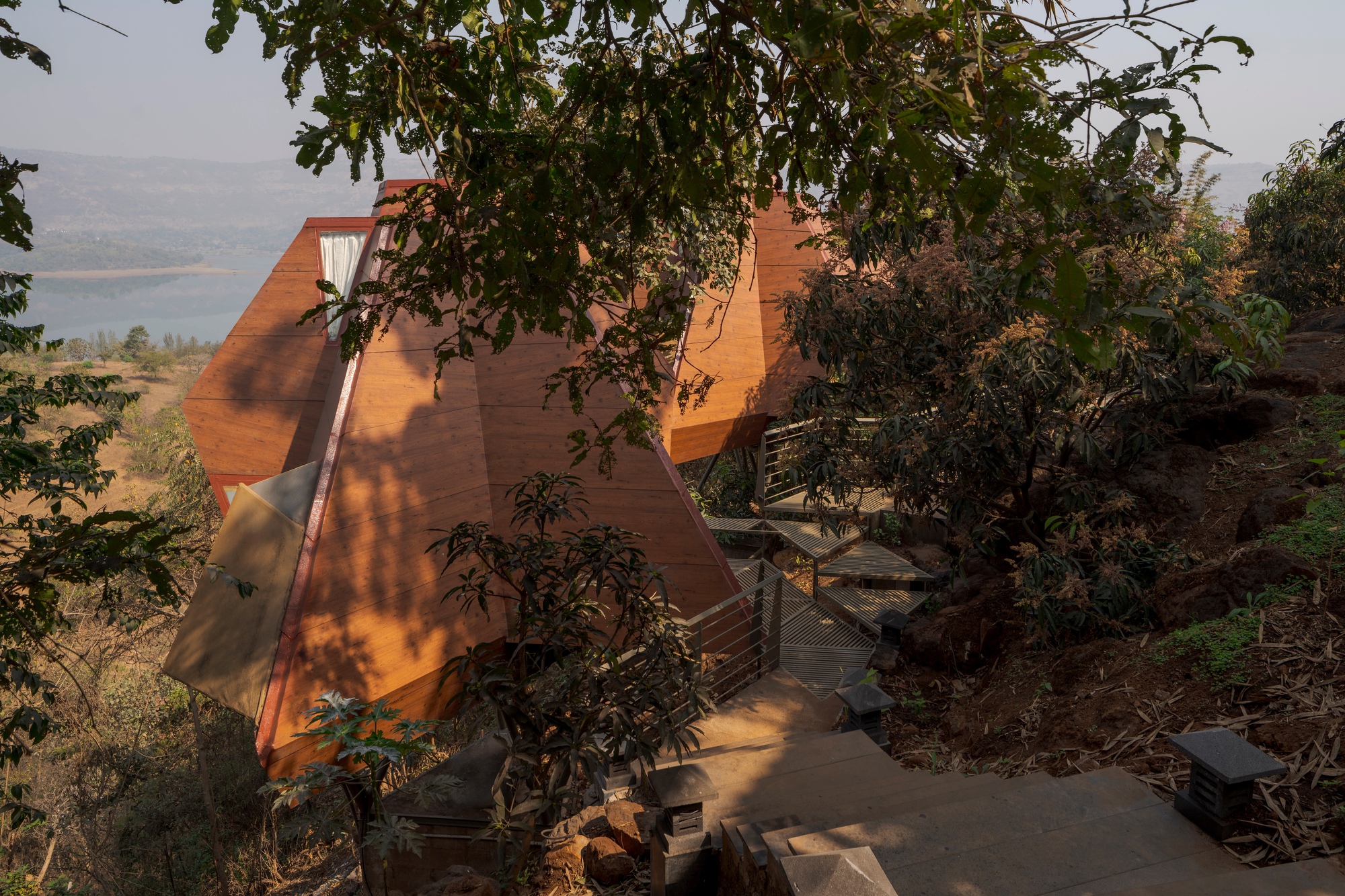 Pyramidal timber structure rising from hillside site with lake visible in distant valley beyond