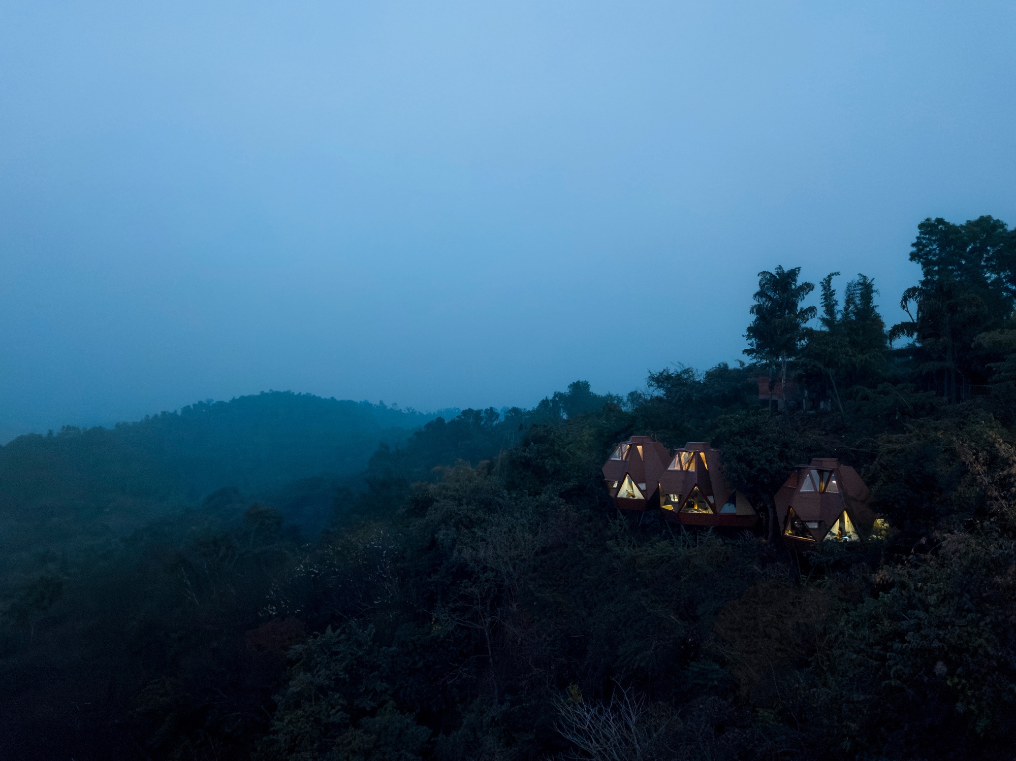 Cluster of peaked pavilion roofs illuminated from within on a forested hillside at dusk