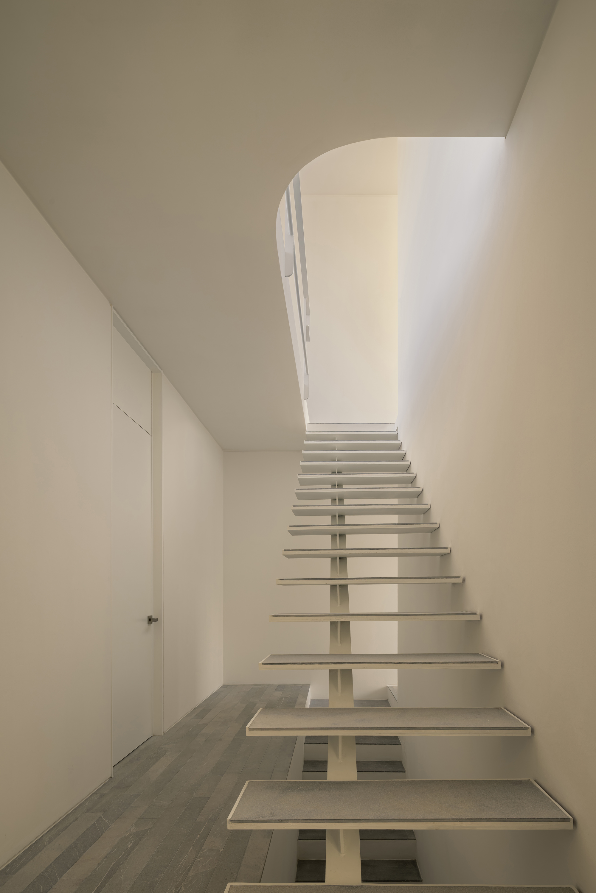 Open-riser staircase with white treads and central spine curving up toward a skylit landing