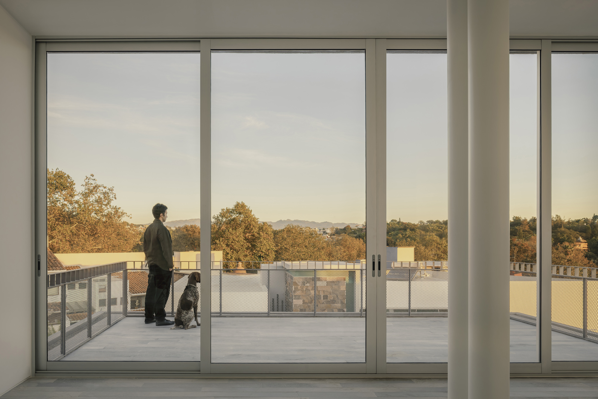 Interior view through full-height glass doors to a rooftop terrace where a person stands with a dog at dusk
