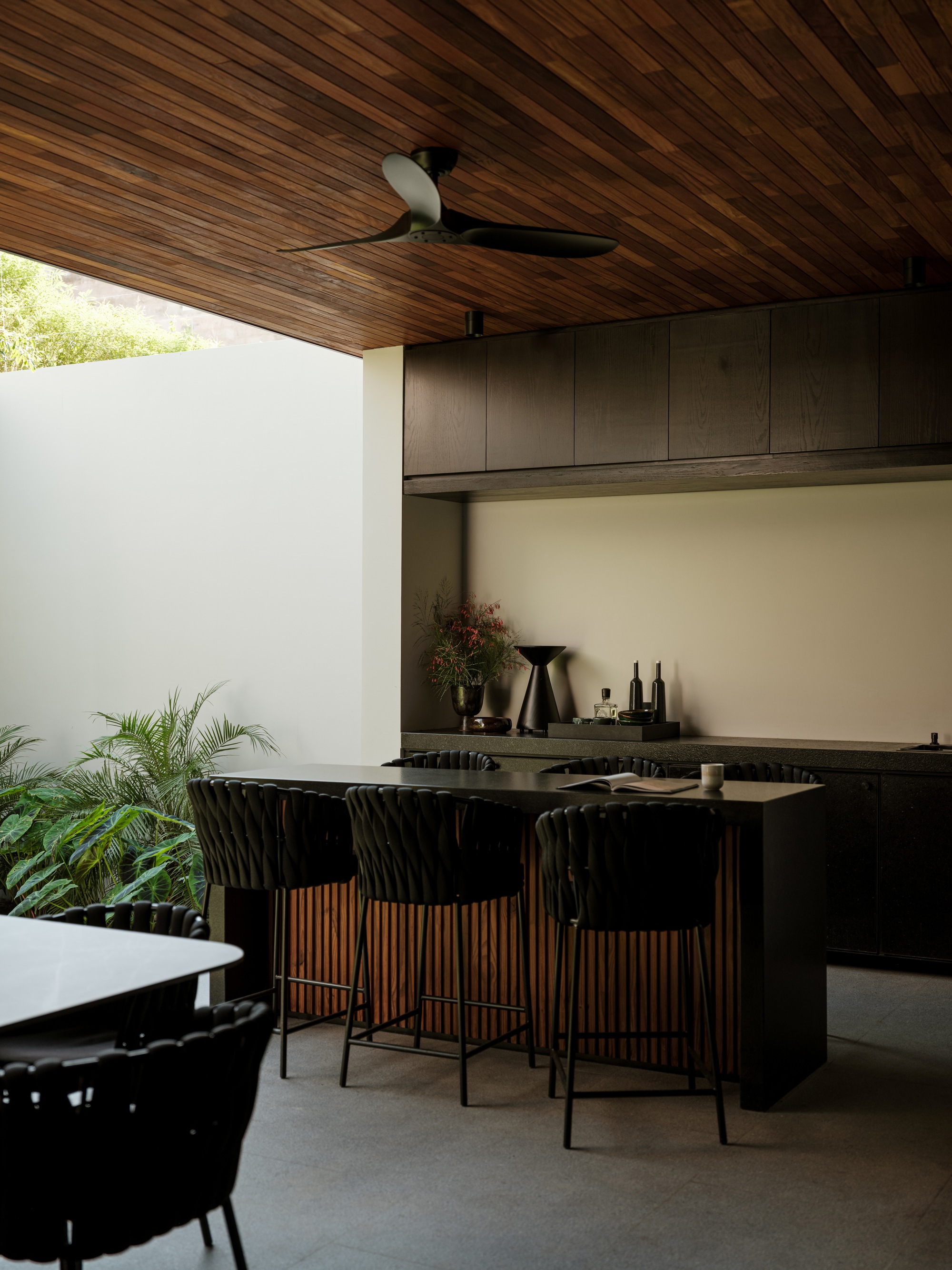 Outdoor kitchen with slatted timber ceiling, black island with bar seating, and tropical greenery beyond