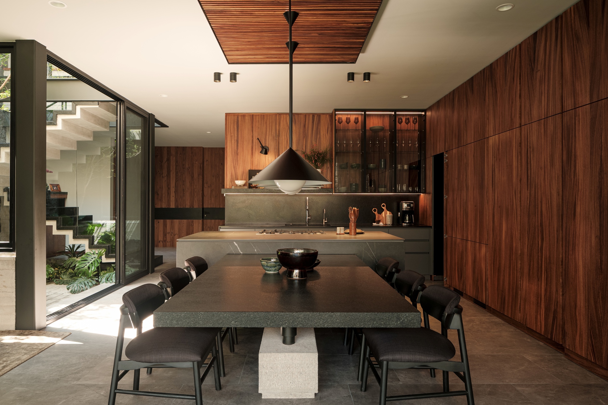 Dining area with timber cabinetry and slat ceiling panels opening to a planted courtyard with stairs