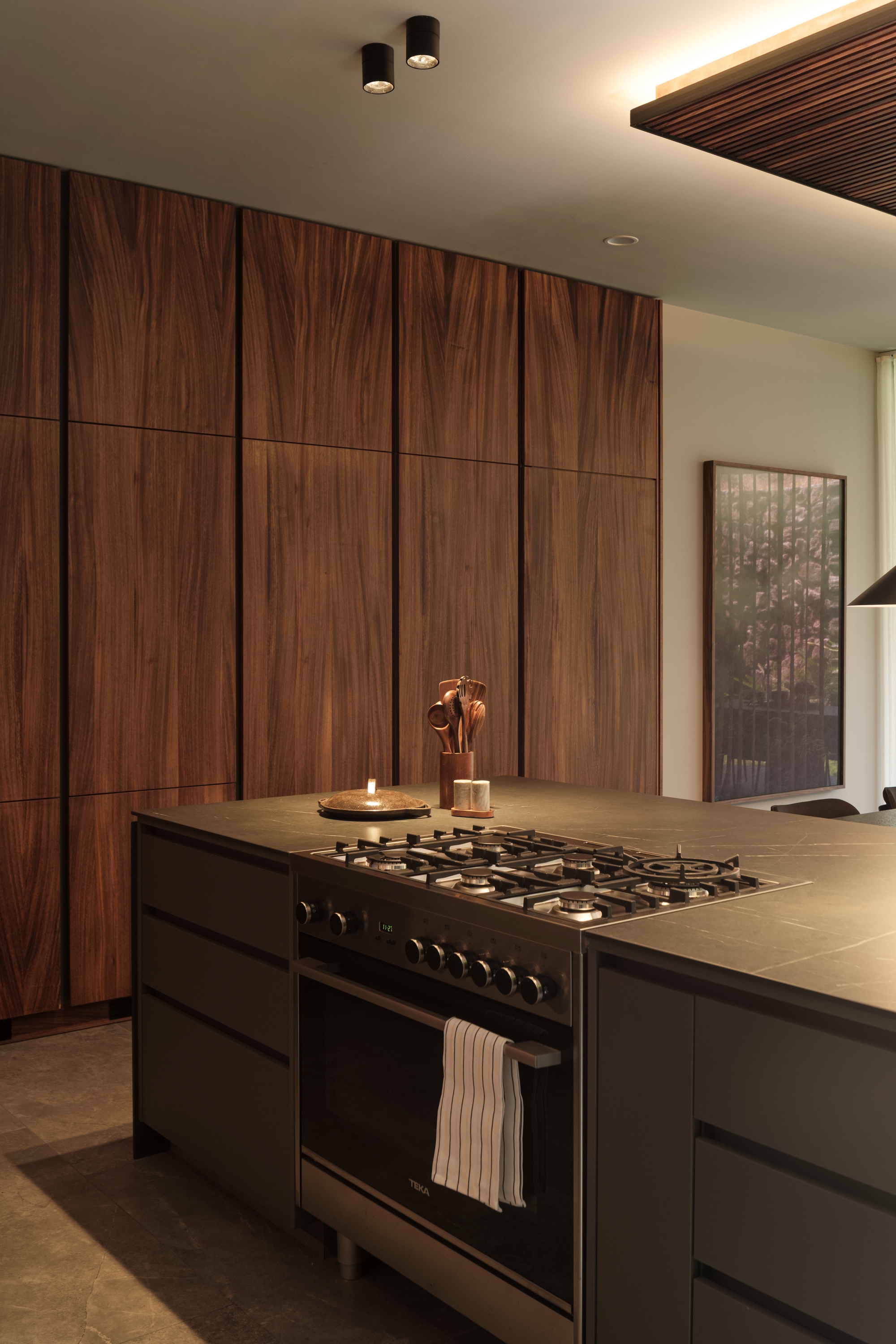 Kitchen island with black gas range against walnut veneer wall beneath recessed cove lighting