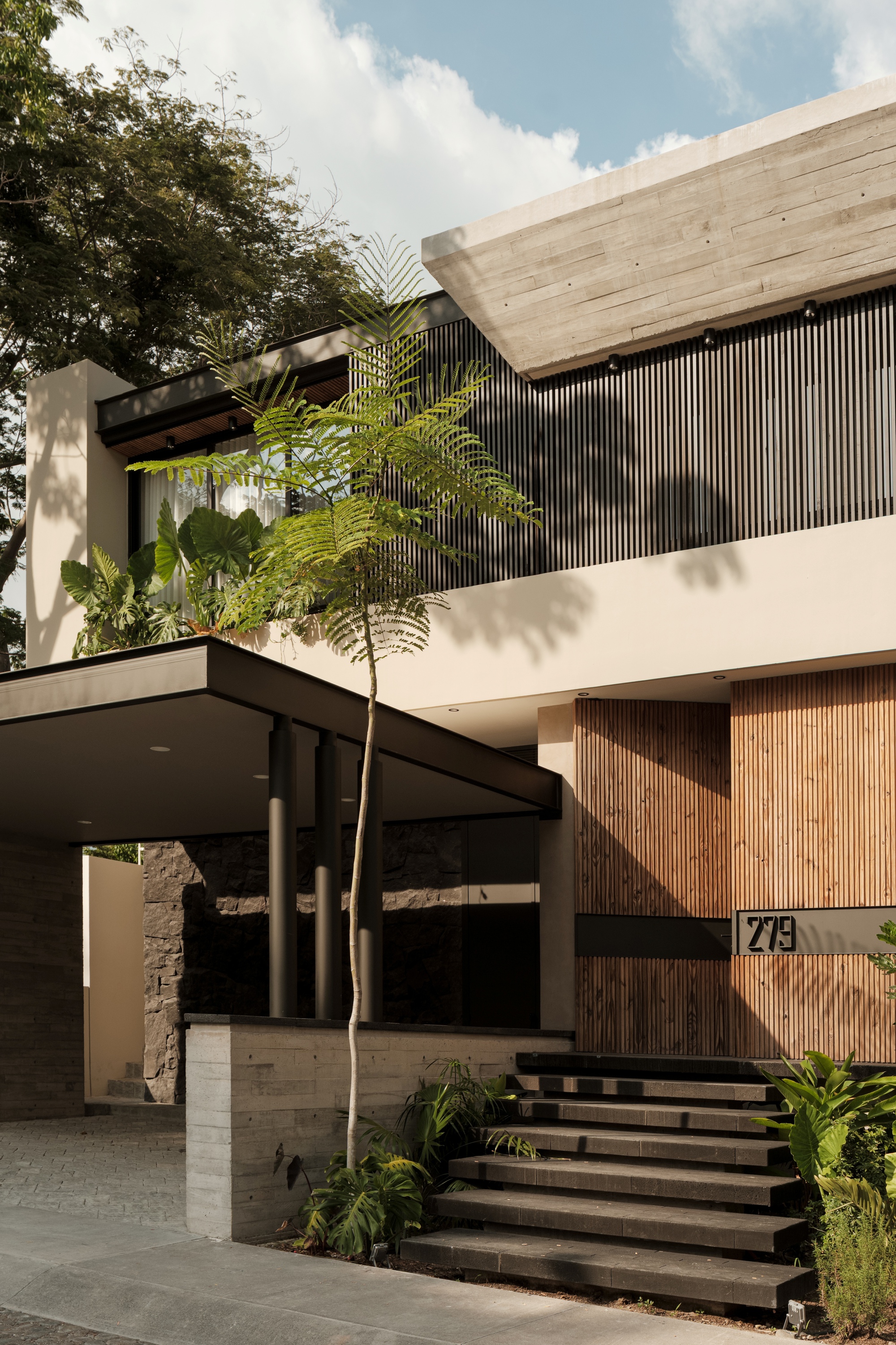 Street facade with vertical wood slat cladding, board-formed concrete canopy, and palm tree shadows