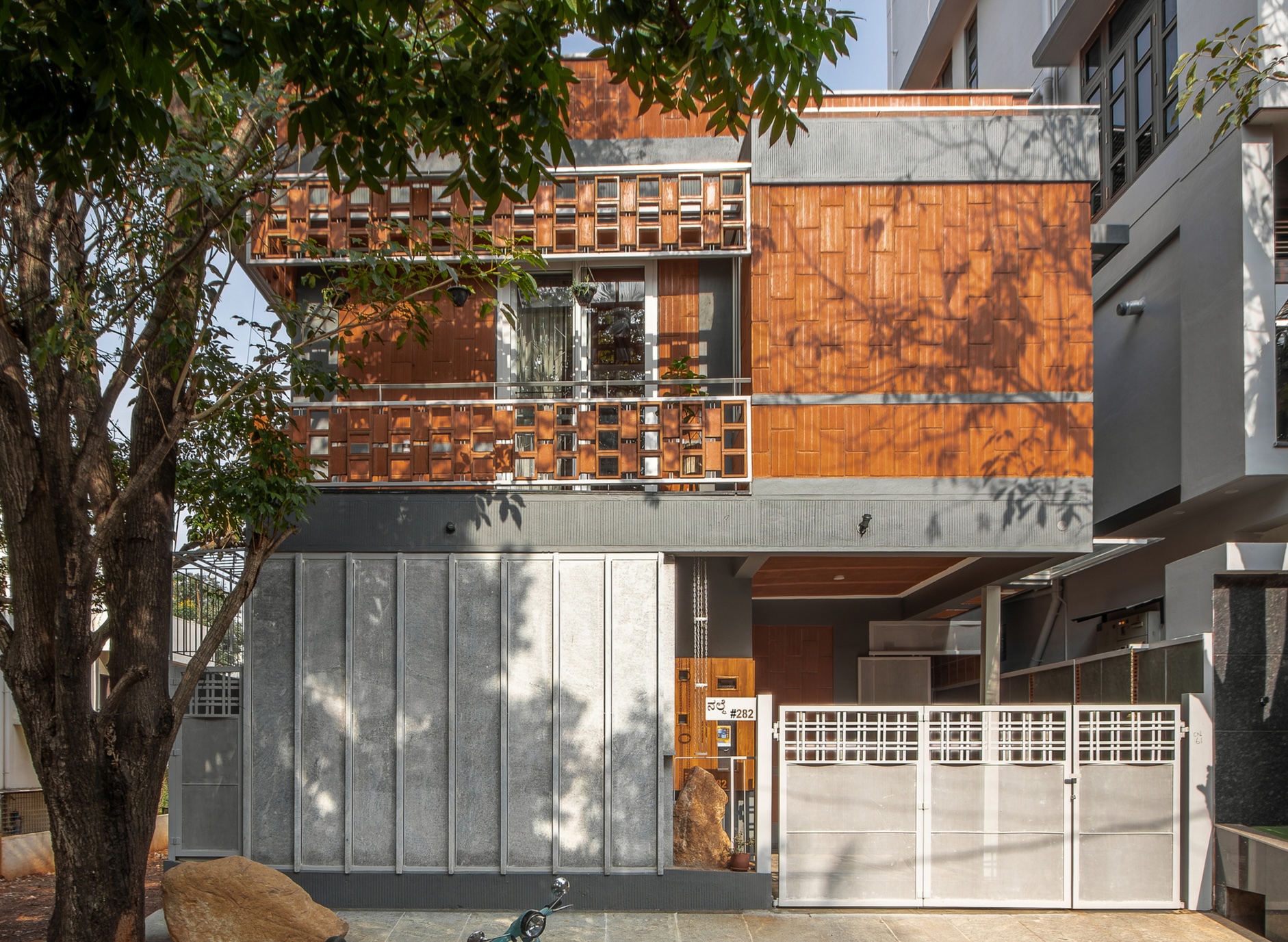 Street facade with orange terracotta tile screens above grey metal panels and dappled tree shadows