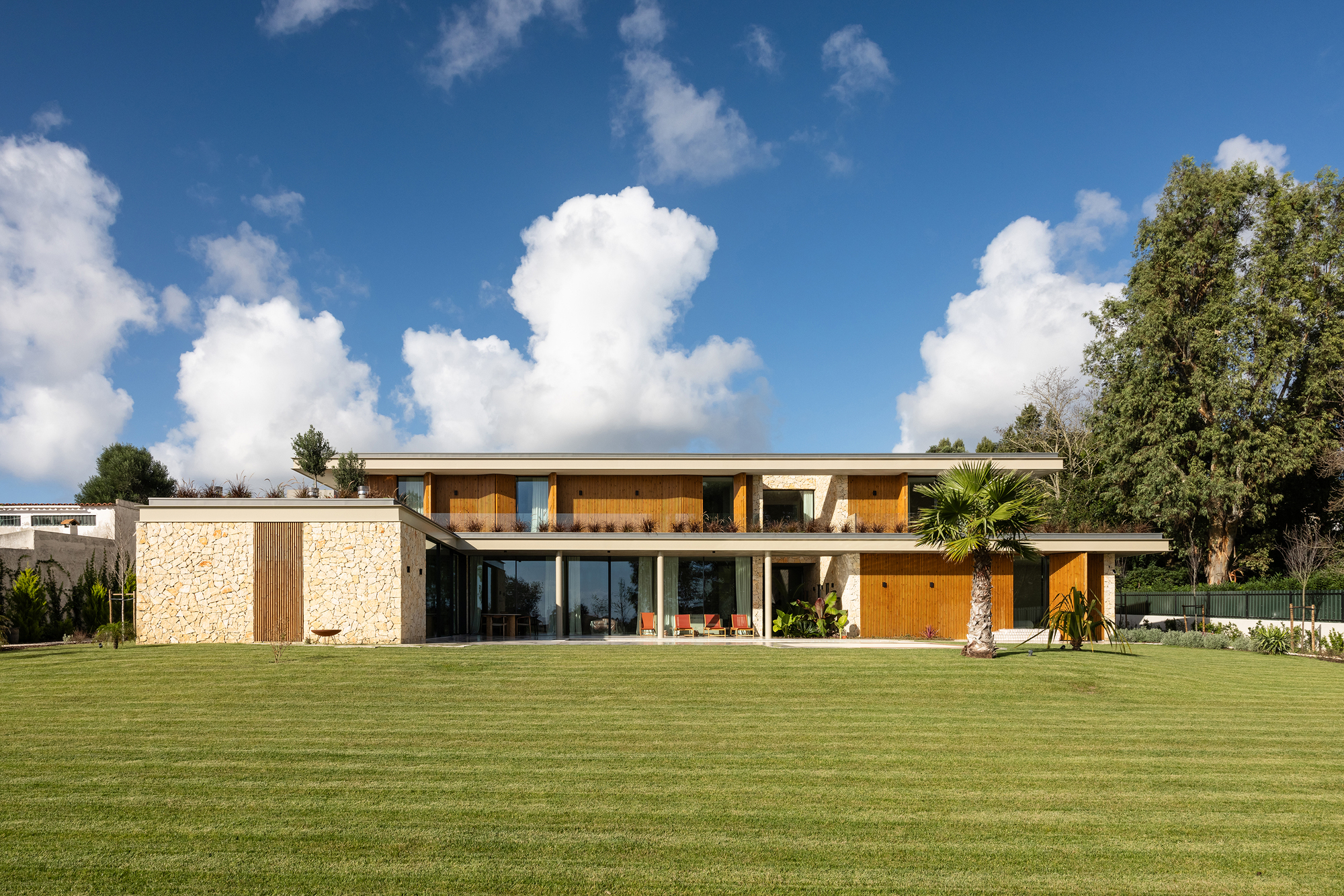 Garden view of the two-storey facade with stone walls, timber screens and expansive glazing under white clouds