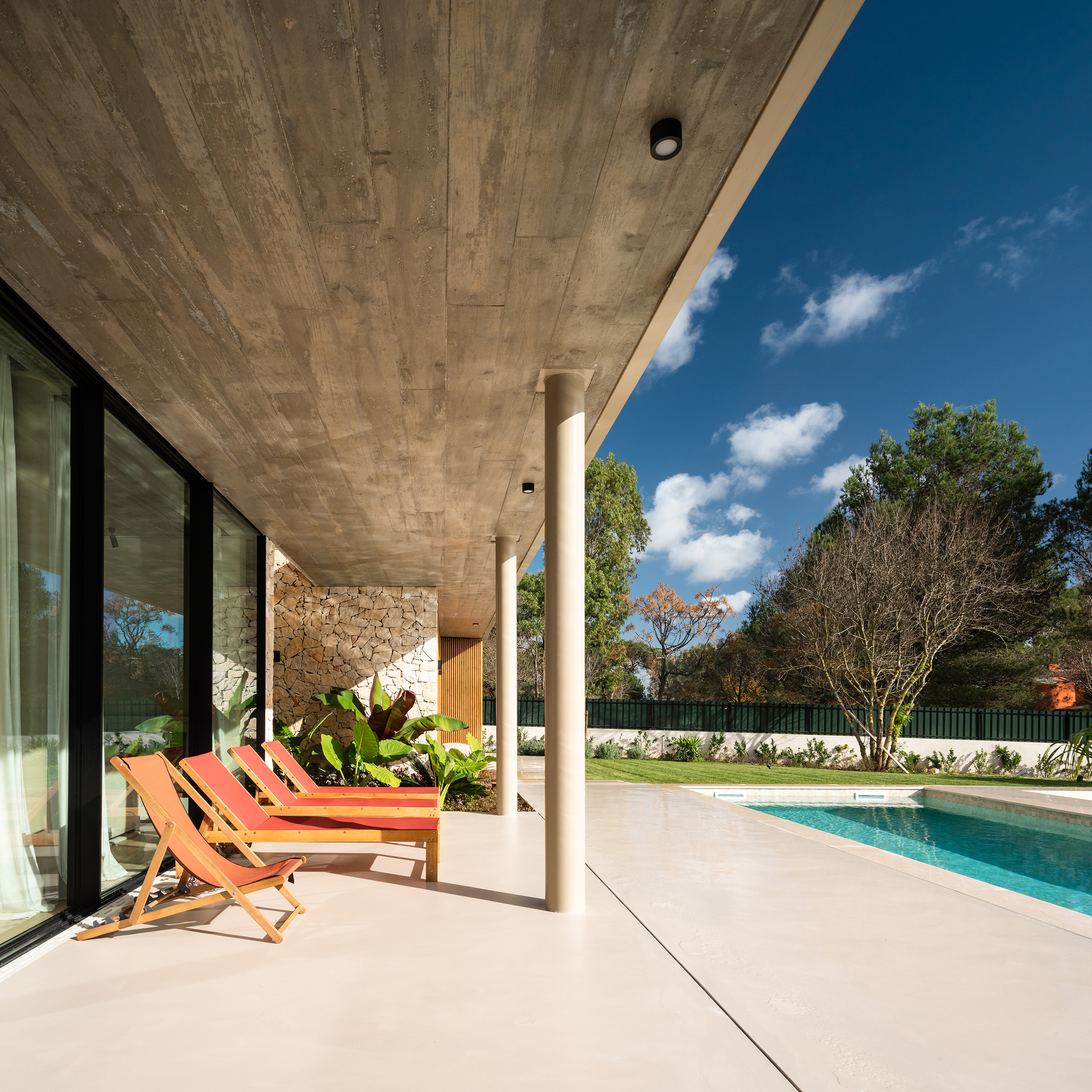 Covered poolside terrace with board-formed concrete soffit and cylindrical white column under blue sky