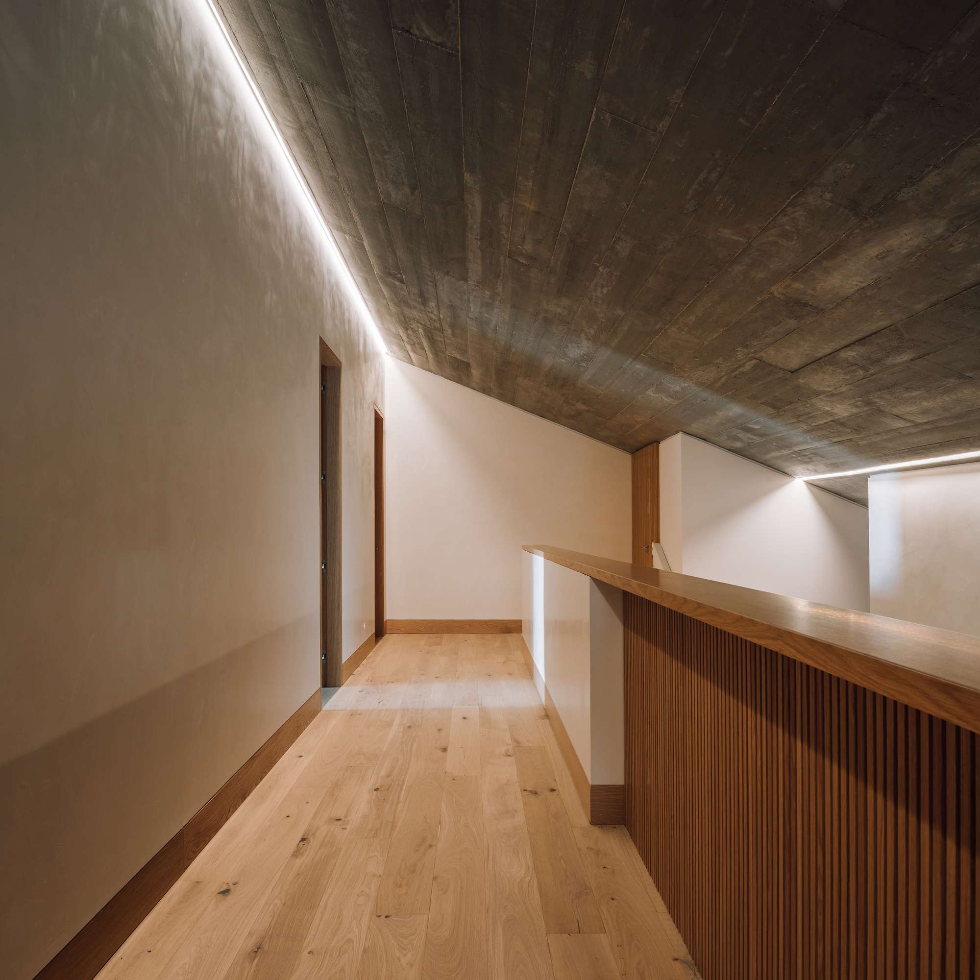 Upper corridor with timber-slatted balustrade and exposed board-formed concrete ceiling under linear skylight