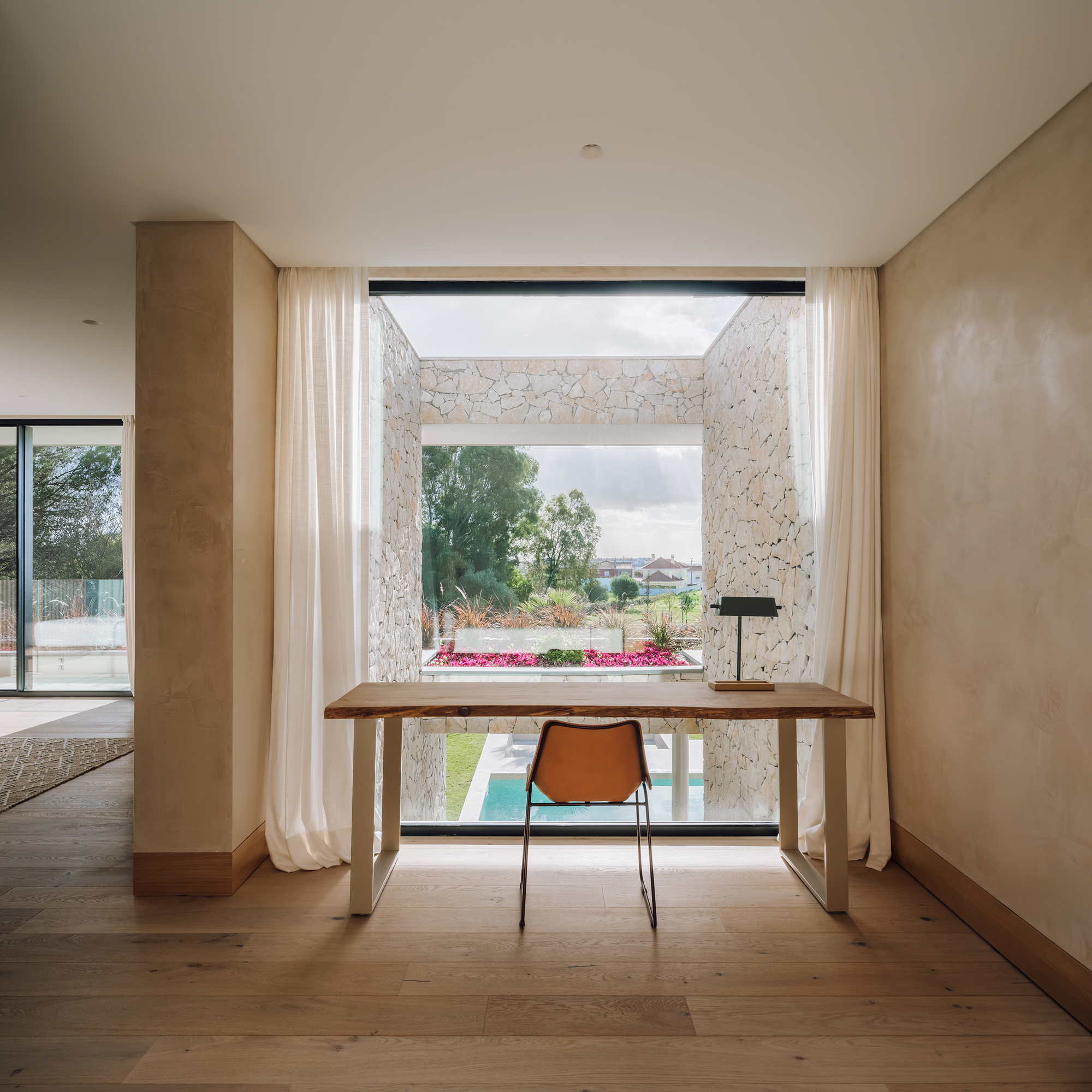 Work desk positioned at full-height window framed by sheer curtains overlooking stone-walled courtyard